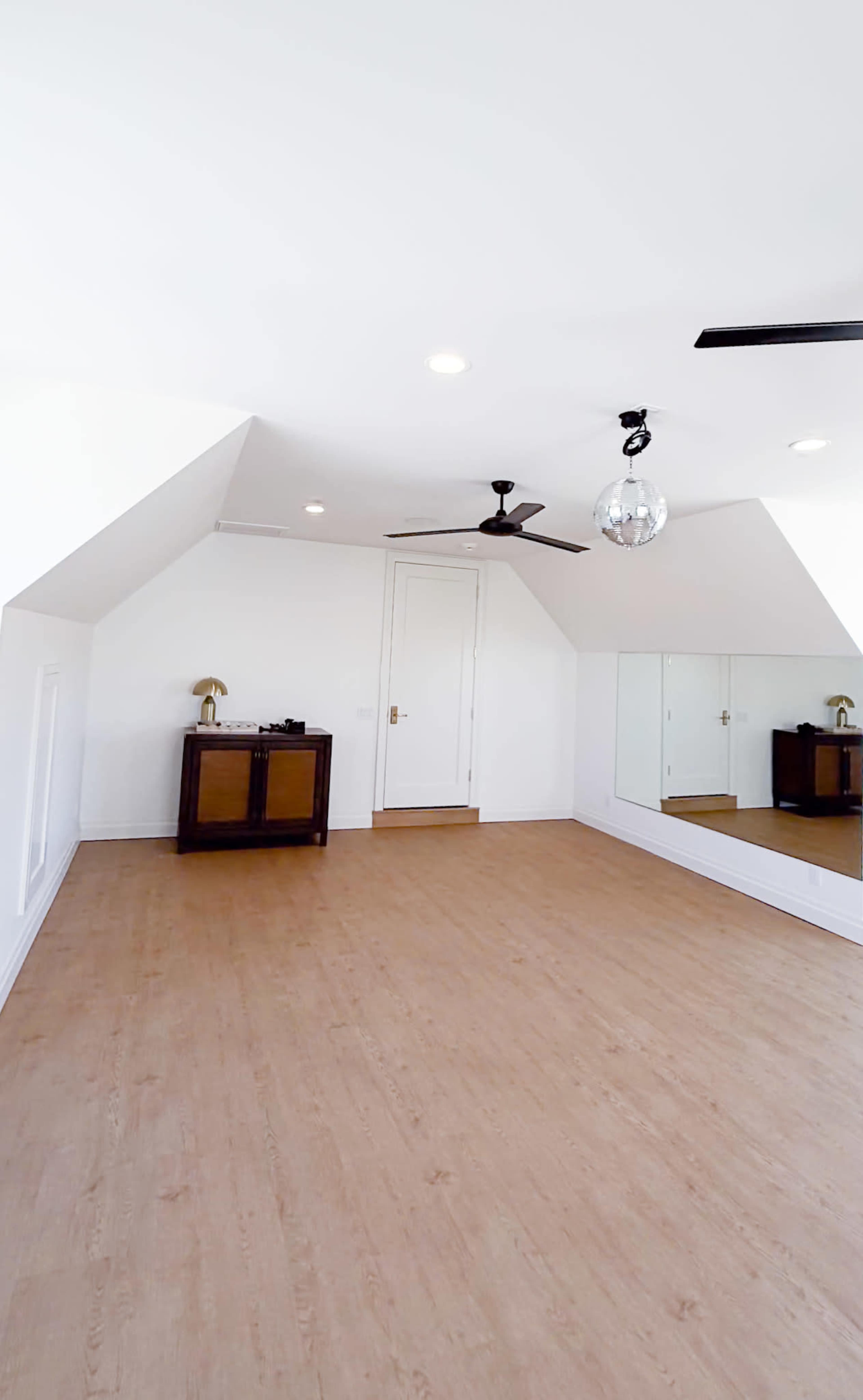 The image shows a spacious, well-lit attic room with wooden flooring, a ceiling fan, a mirror wall, and a vintage cabinet against one wall.