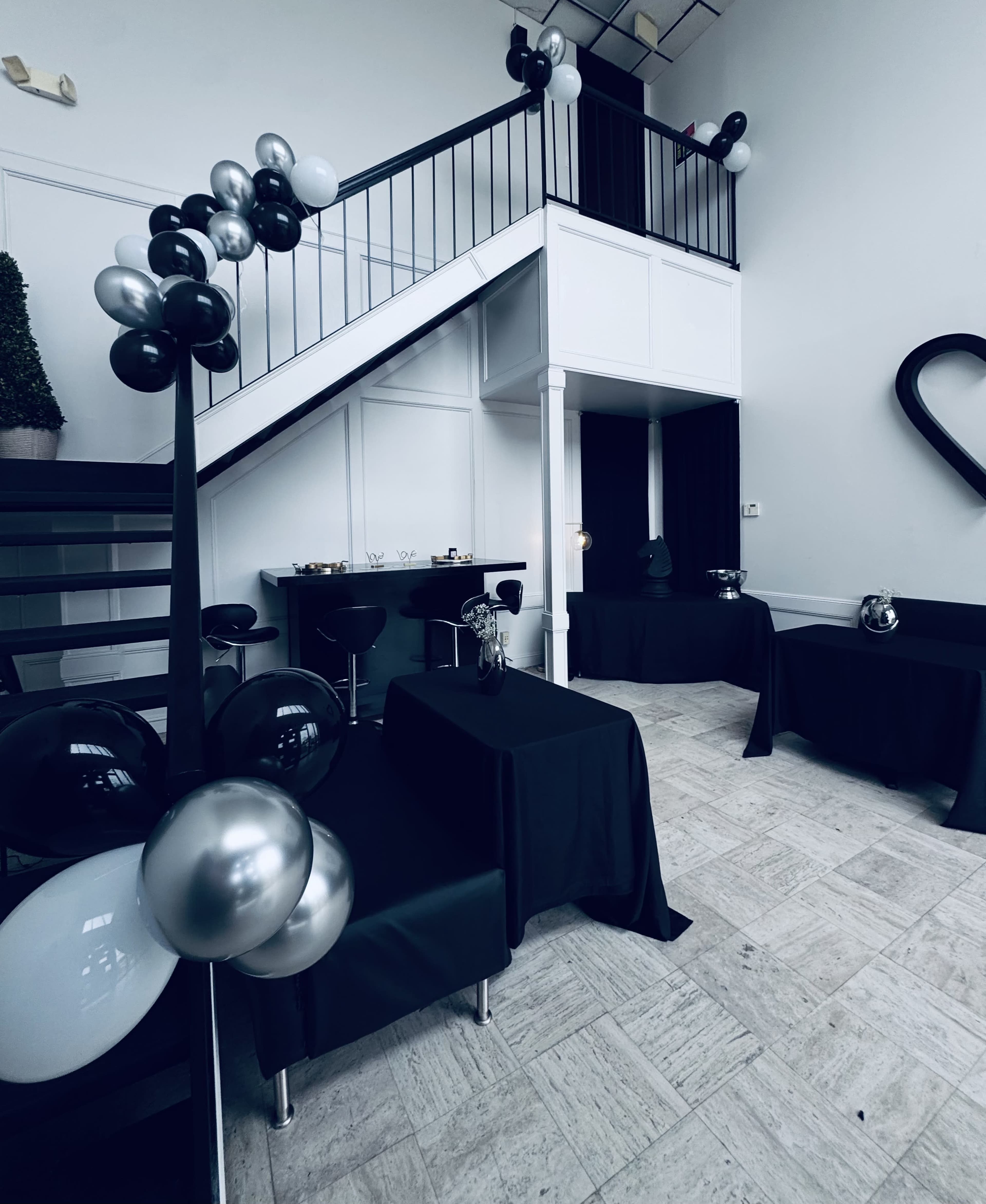 The image shows a spacious event space featuring black and white decorations, a staircase, and tables covered in black cloth.