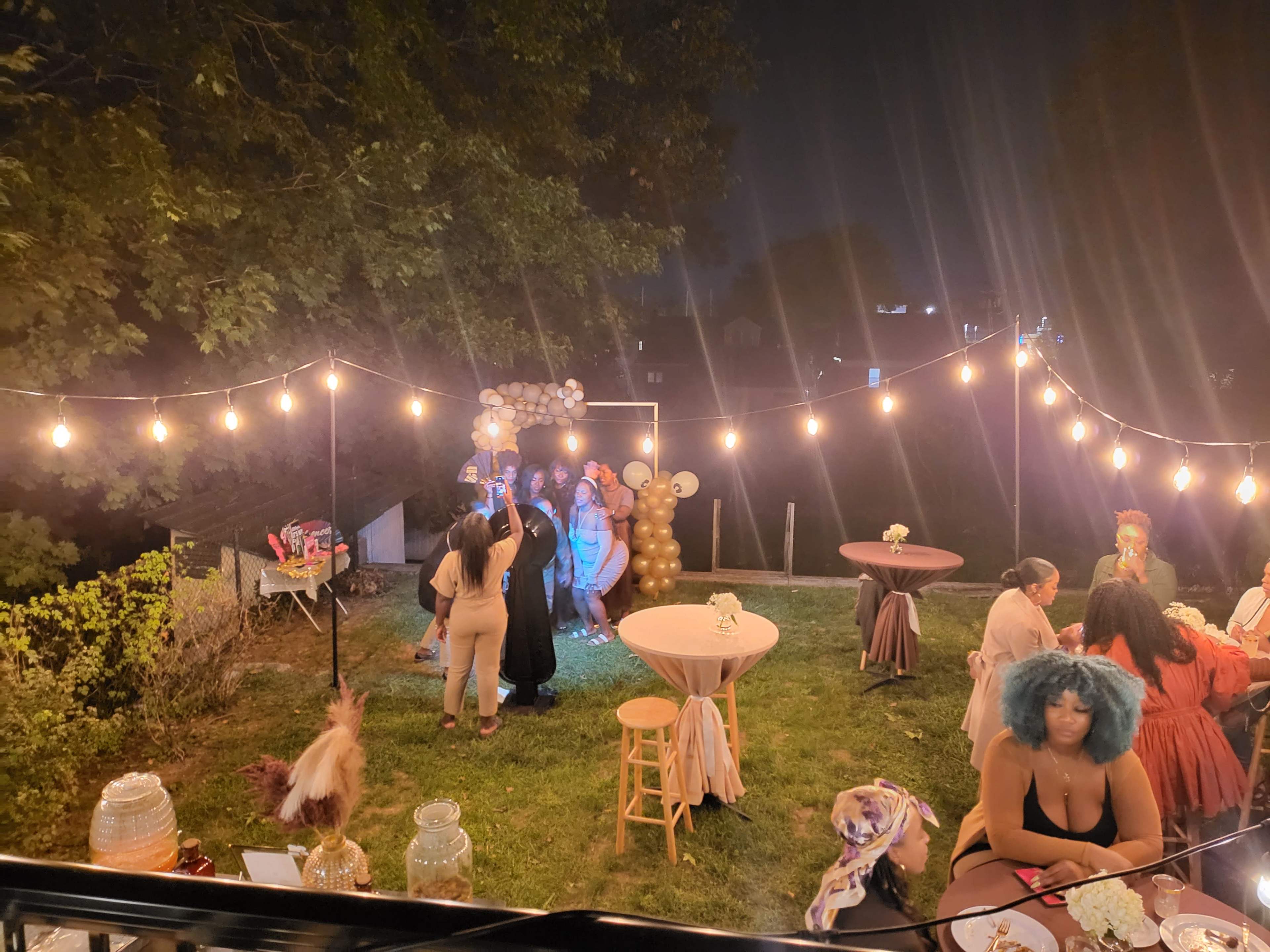 A garden party is illuminated by string lights, featuring guests dancing and dining among decorated tables.