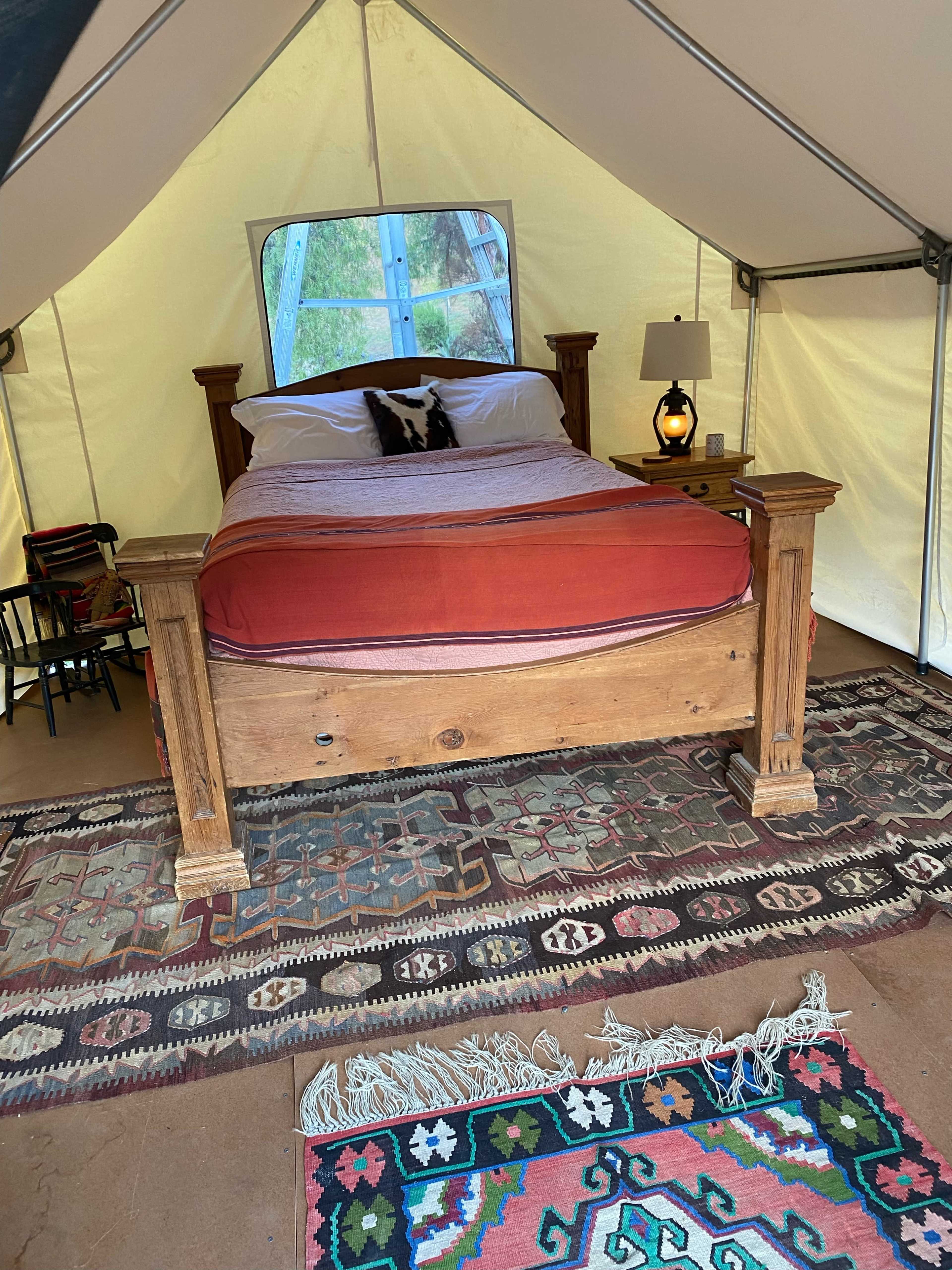 A tent interior featuring a wooden bed with red bedding, a nightstand with a lamp, and decorative rugs on a floor.