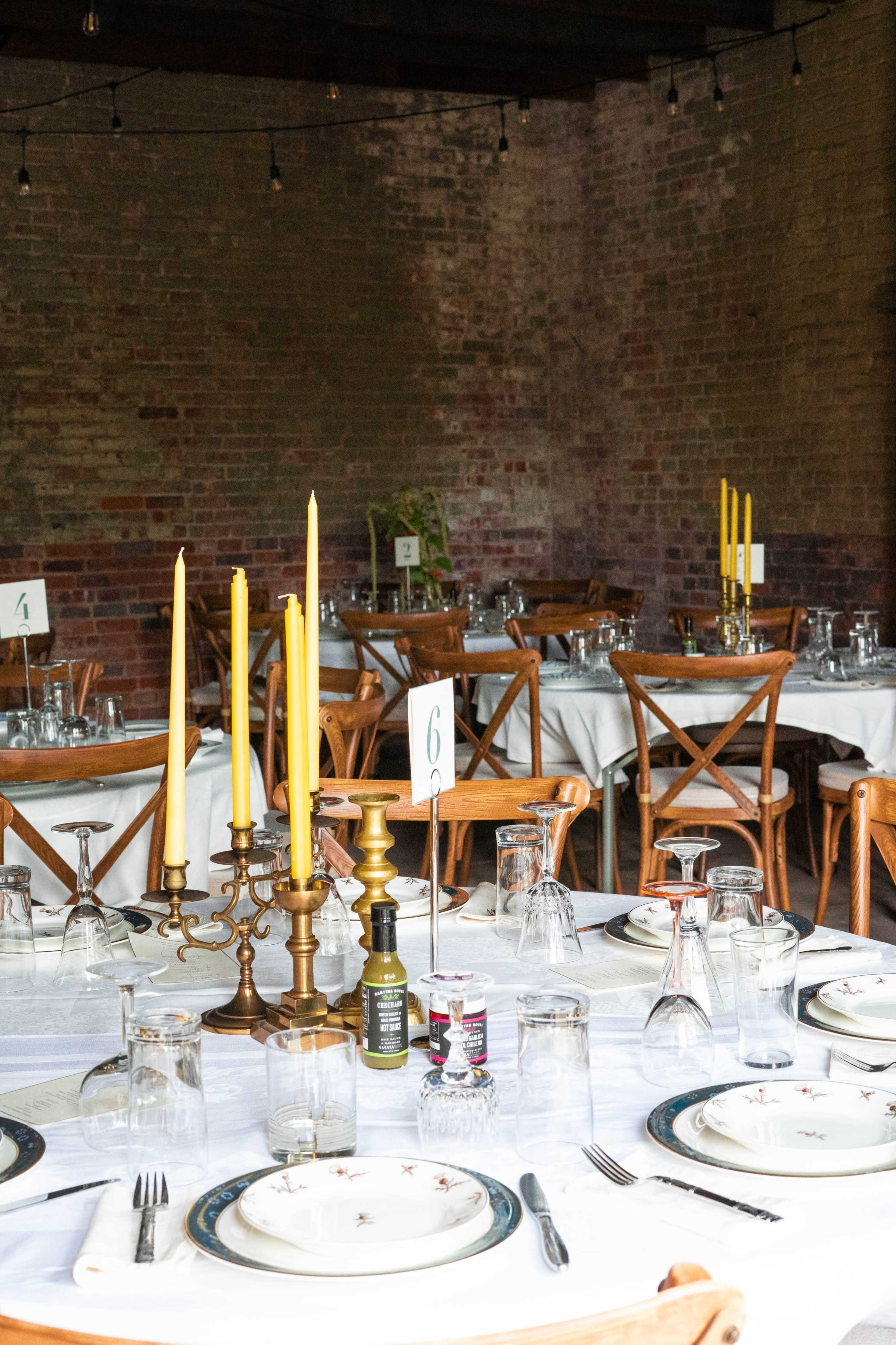 The image shows a dining setup with several tables arranged in a room featuring exposed brick walls, each table adorned with white tablecloths, plates, glasses, and decorative candles.