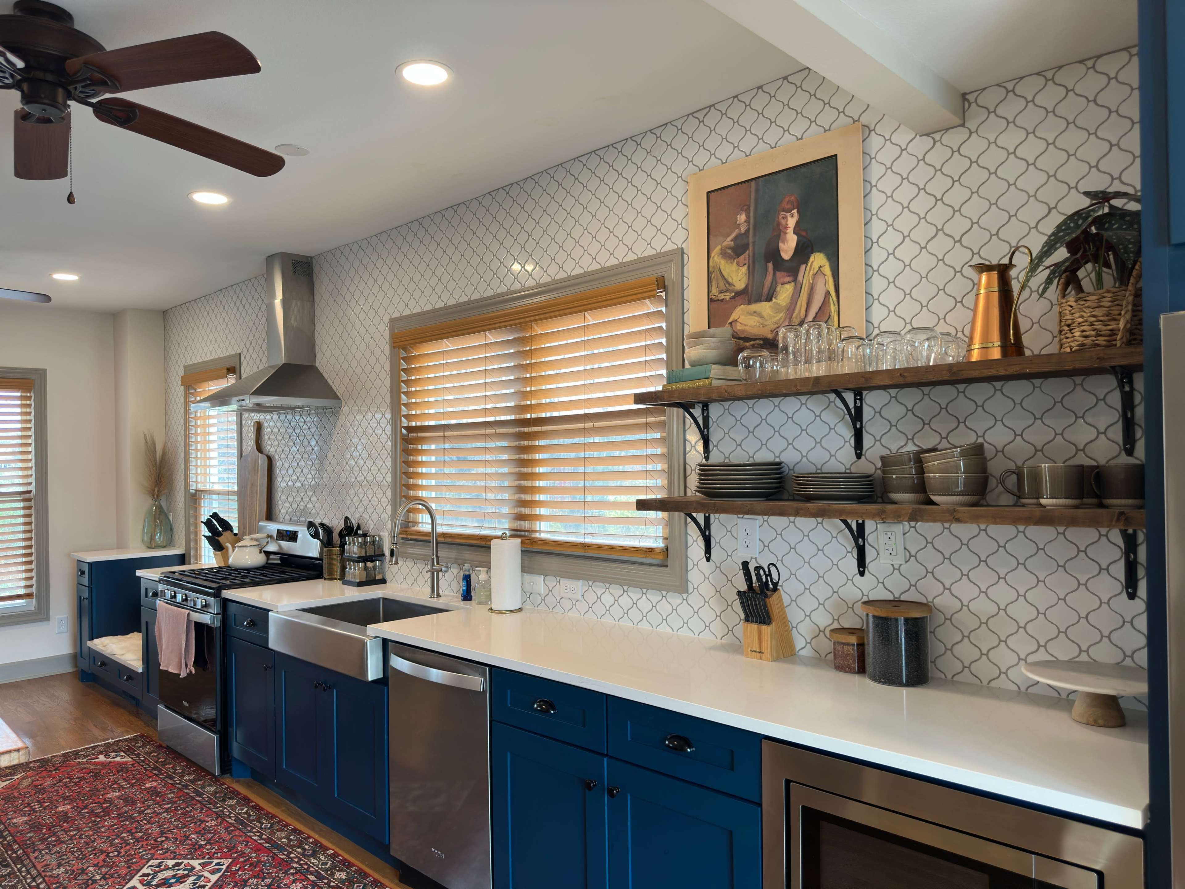 The image shows a modern kitchen featuring blue cabinetry, a stainless steel stove, and open shelves displaying dishes and glassware against a patterned tile backsplash.