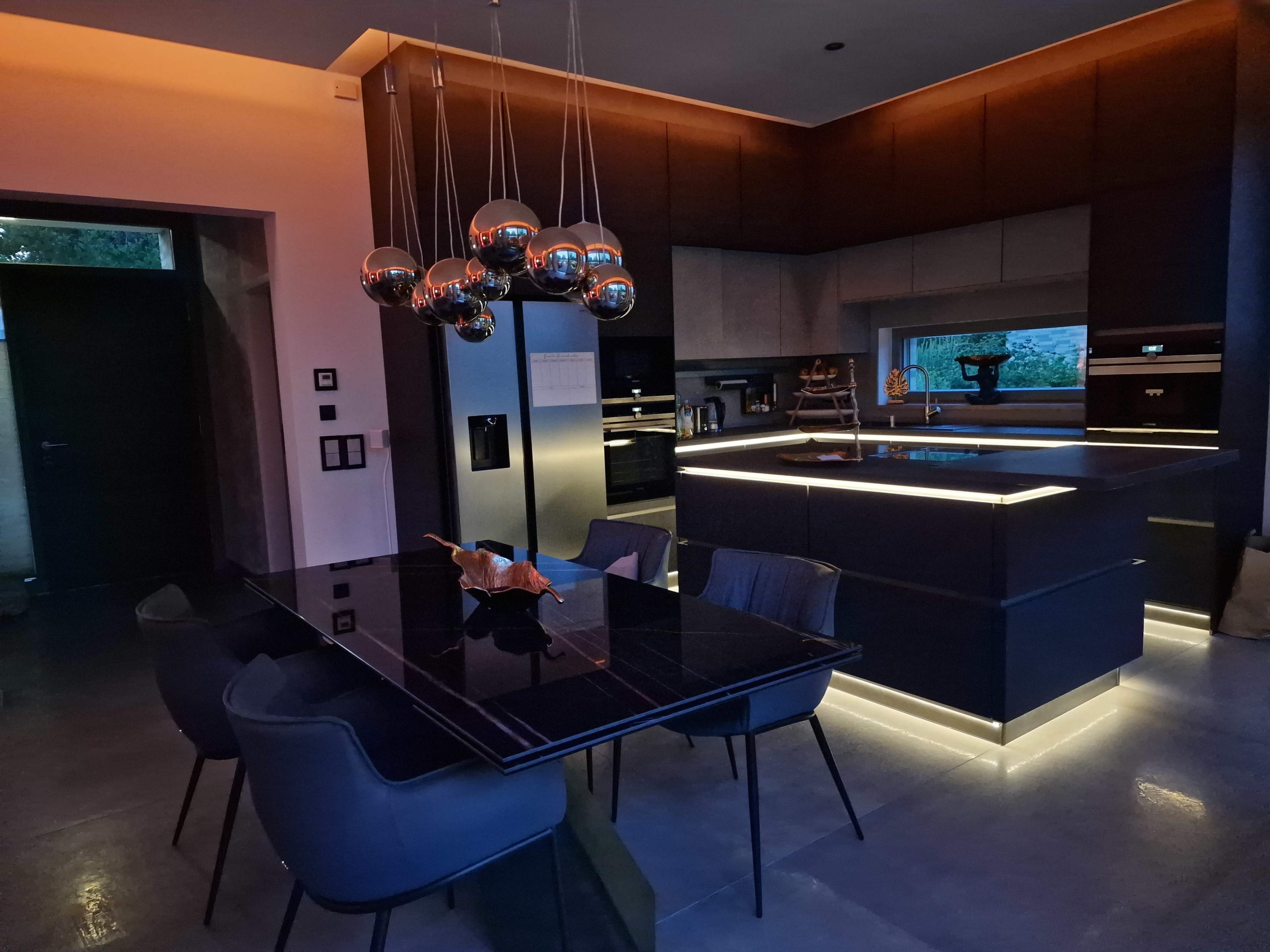A modern kitchen and dining area featuring a dark table, stylish chairs, and pendant lighting over a kitchen island with backlit cabinetry.