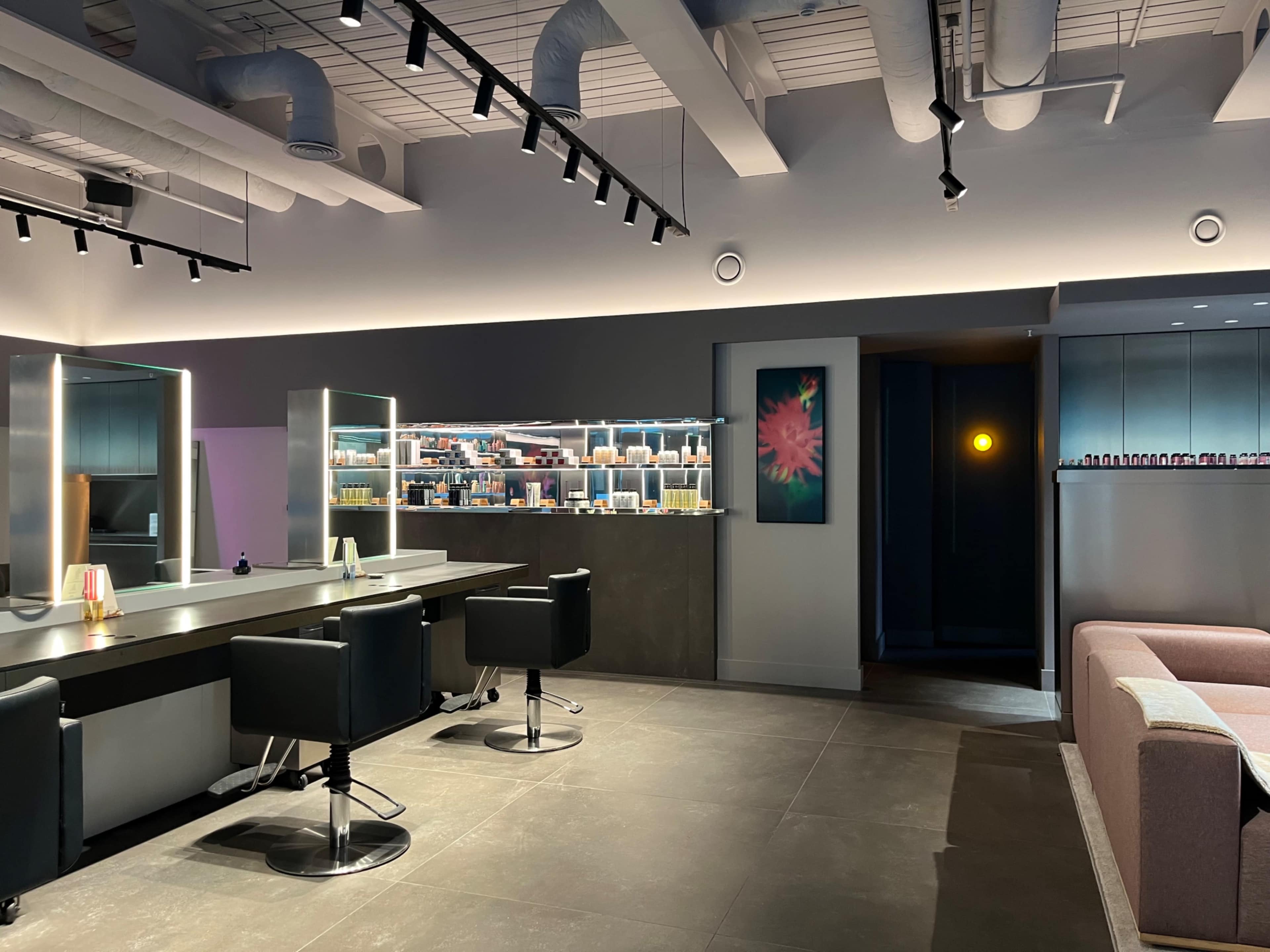 The image shows a modern salon interior with styling chairs, a product display shelf, and a cozy seating area.