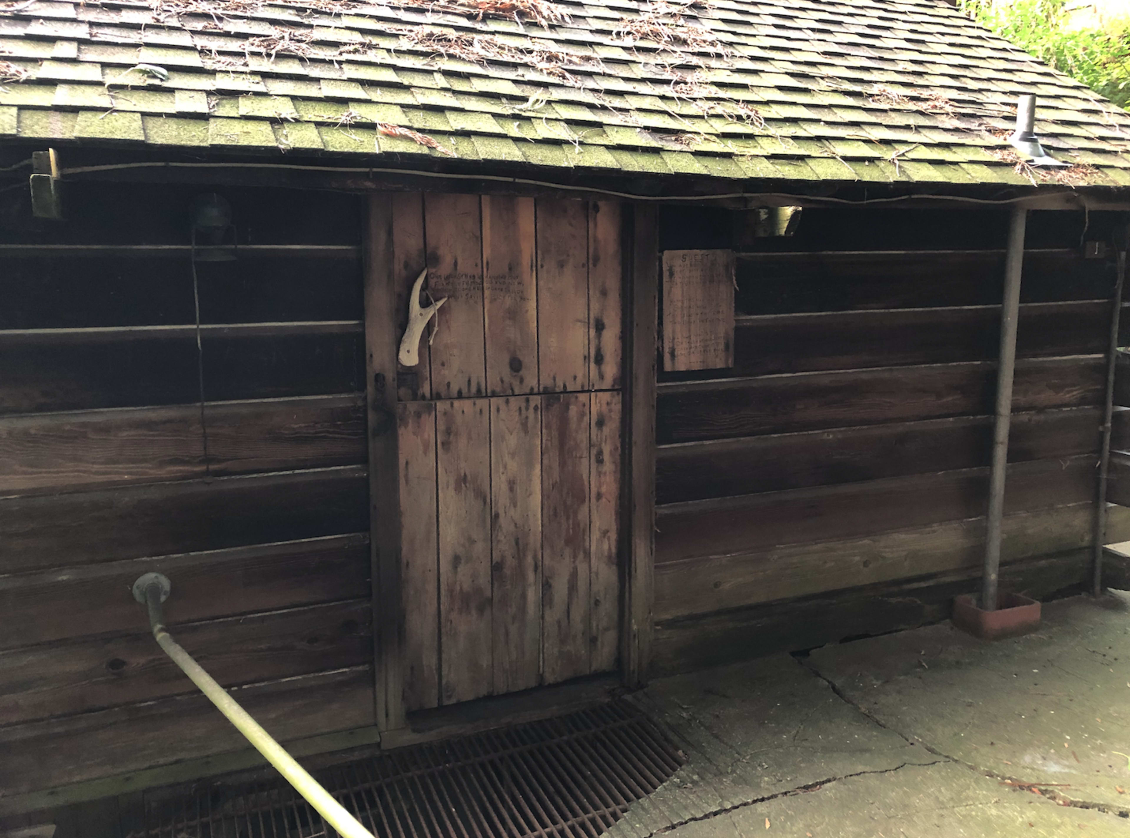 The image shows a wooden cabin with a sloped roof, featuring a door with a rustic appearance and a few small items attached nearby.