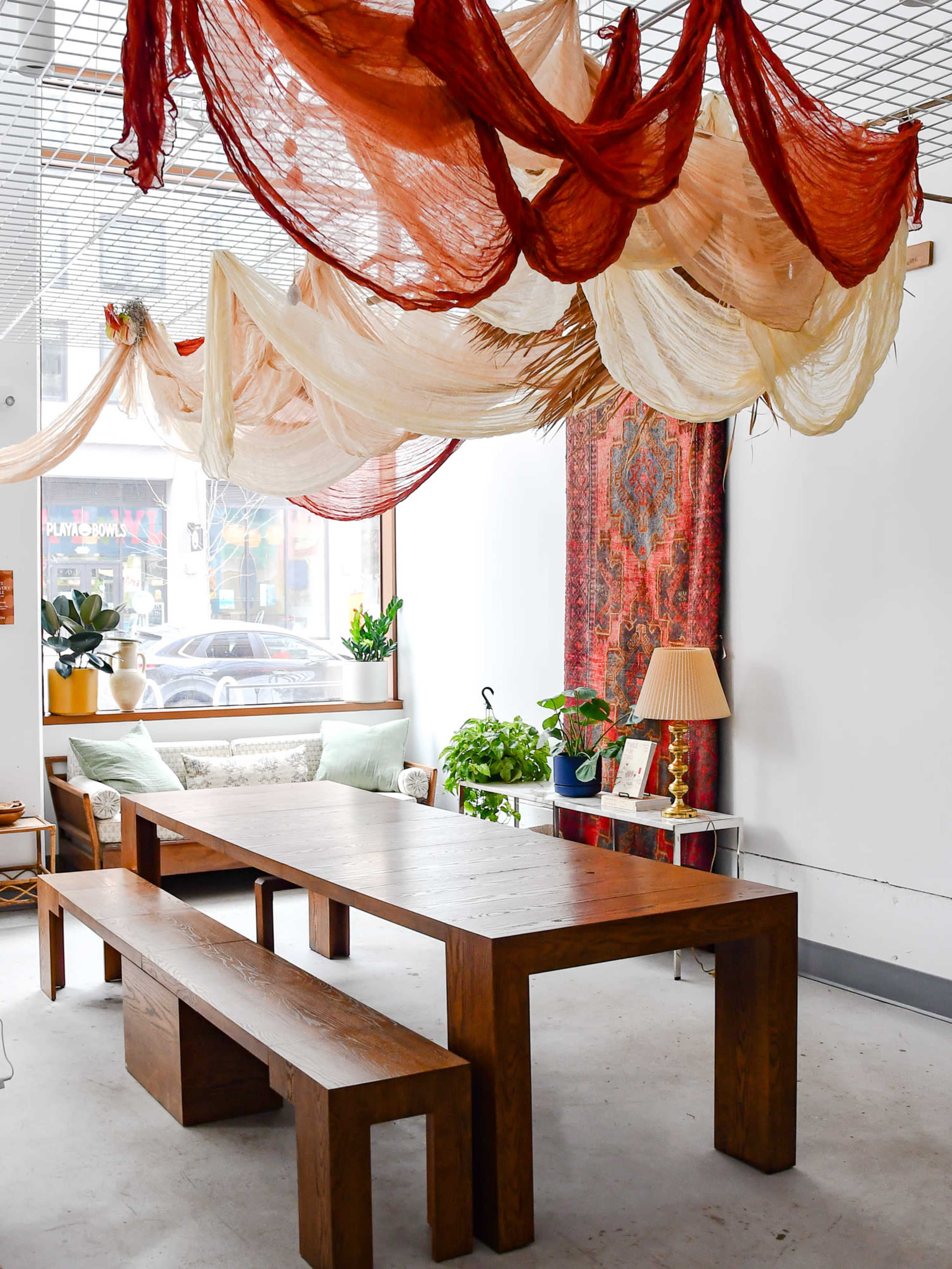 The image shows a cozy interior space featuring a large wooden table and benches, with colorful fabric draped from the ceiling and plants positioned around the room.