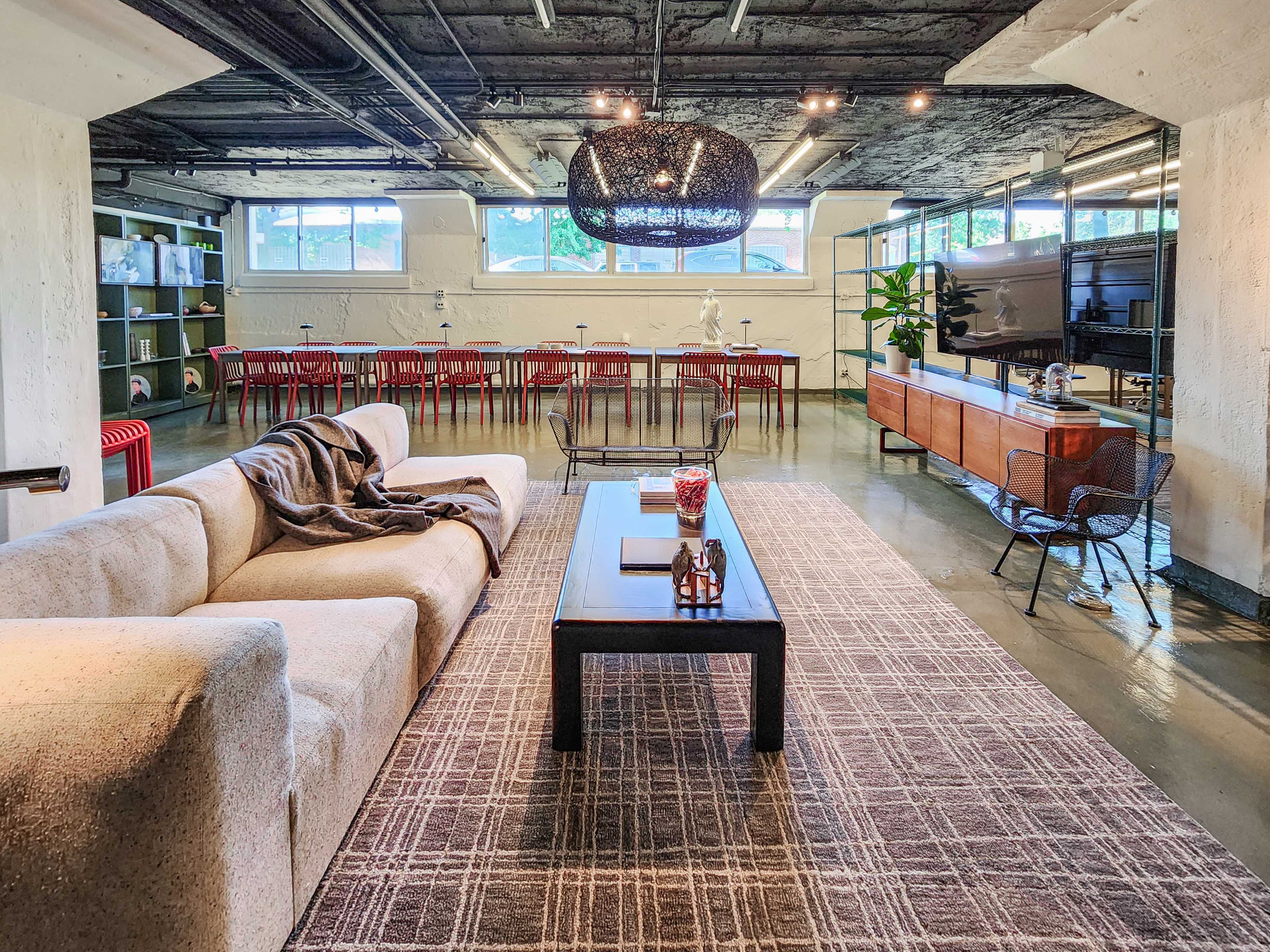 The image shows a modern, open-concept workspace featuring a large beige sofa, a black coffee table, and a dining area with wooden tables and red chairs.