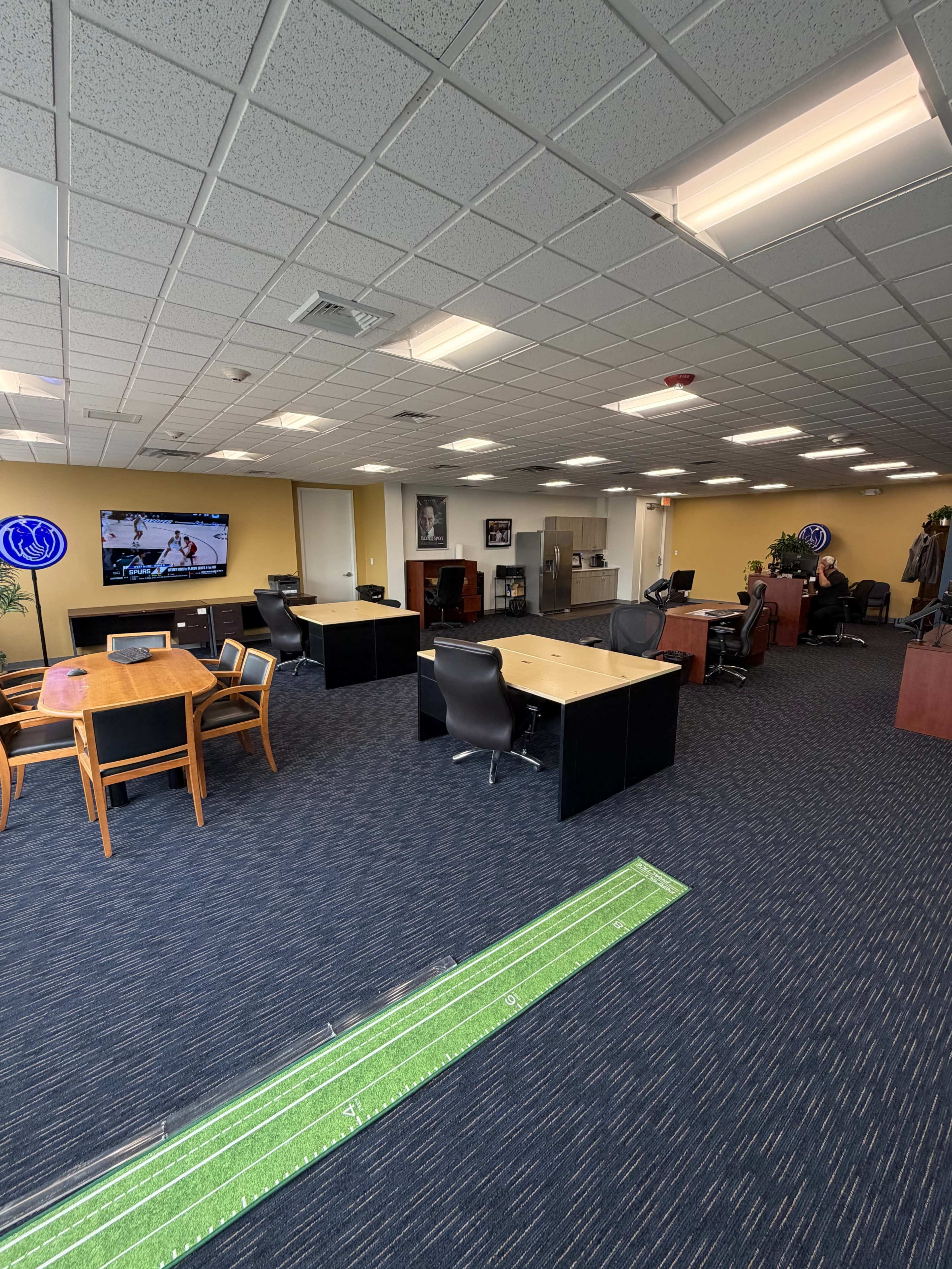 The image depicts a spacious office environment featuring multiple desks, a television, a kitchenette area, and a putting green on the floor.