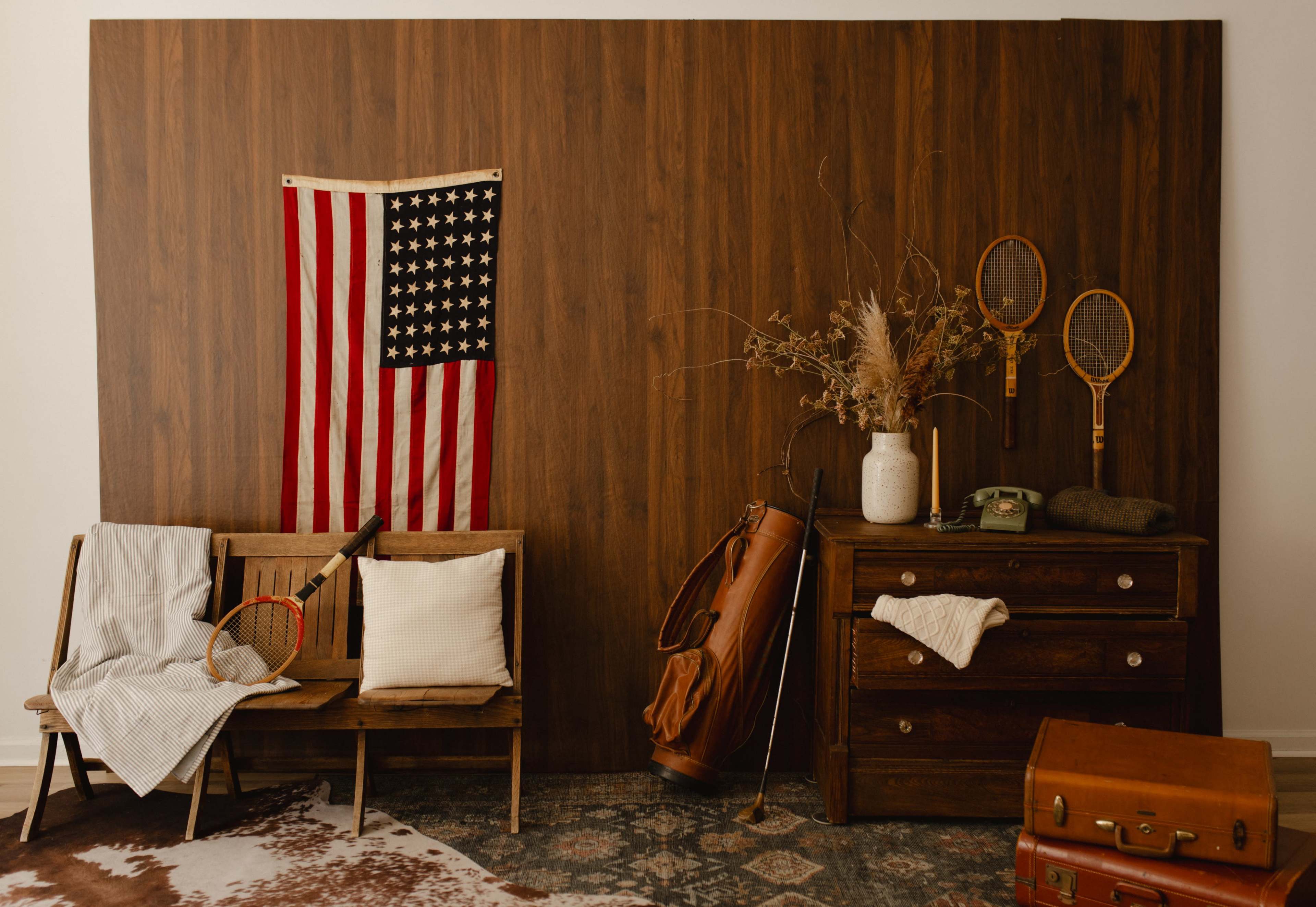 The image features a rustic indoor scene with a wooden wall, an American flag, a vintage tennis racket, a cream-colored vase with dried flowers, a wooden dresser, and various sports equipment arranged artistically.