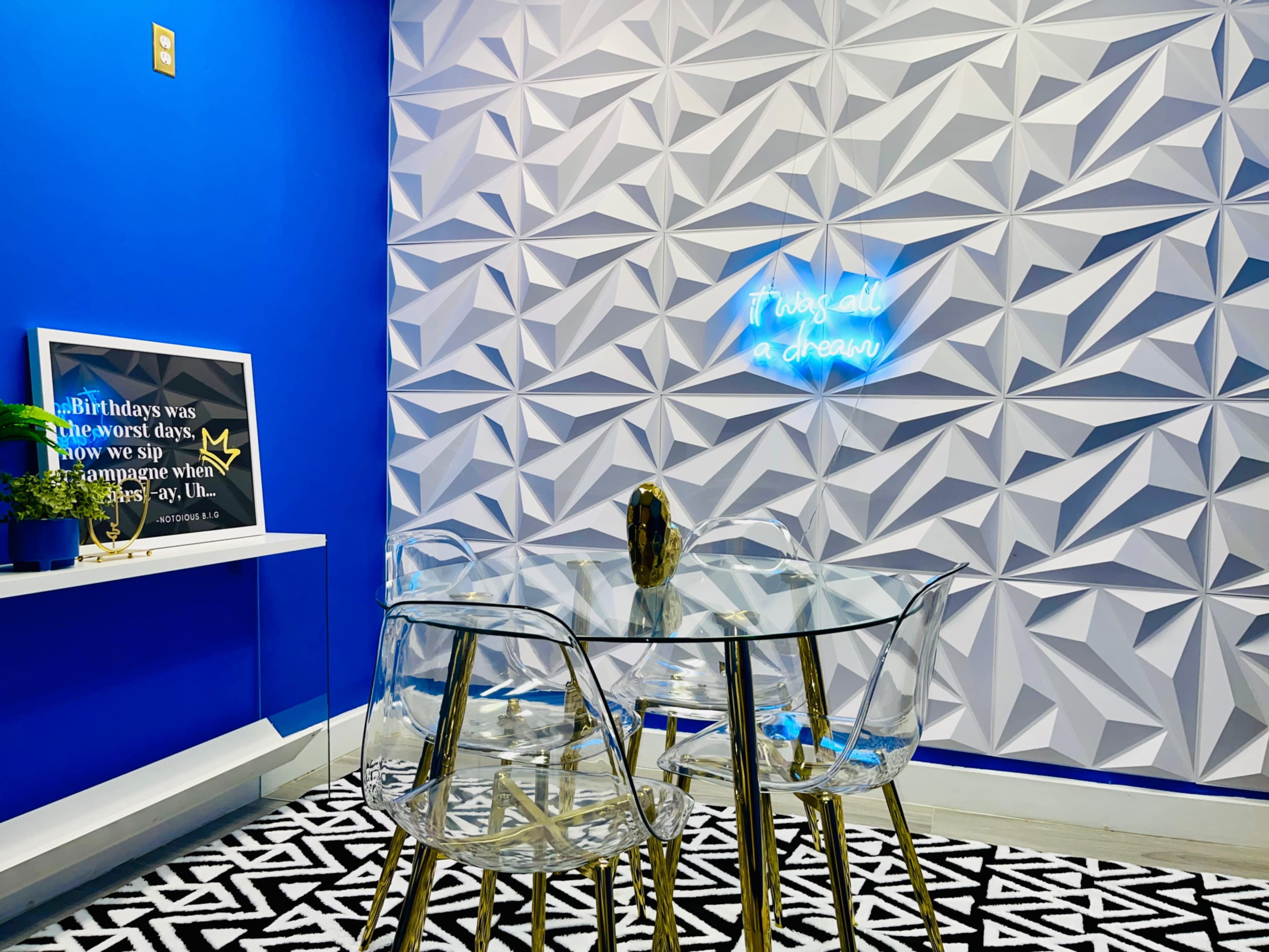 A glass table with transparent chairs is positioned against a textured blue and white wall featuring a neon sign that reads, "It was all a dream."