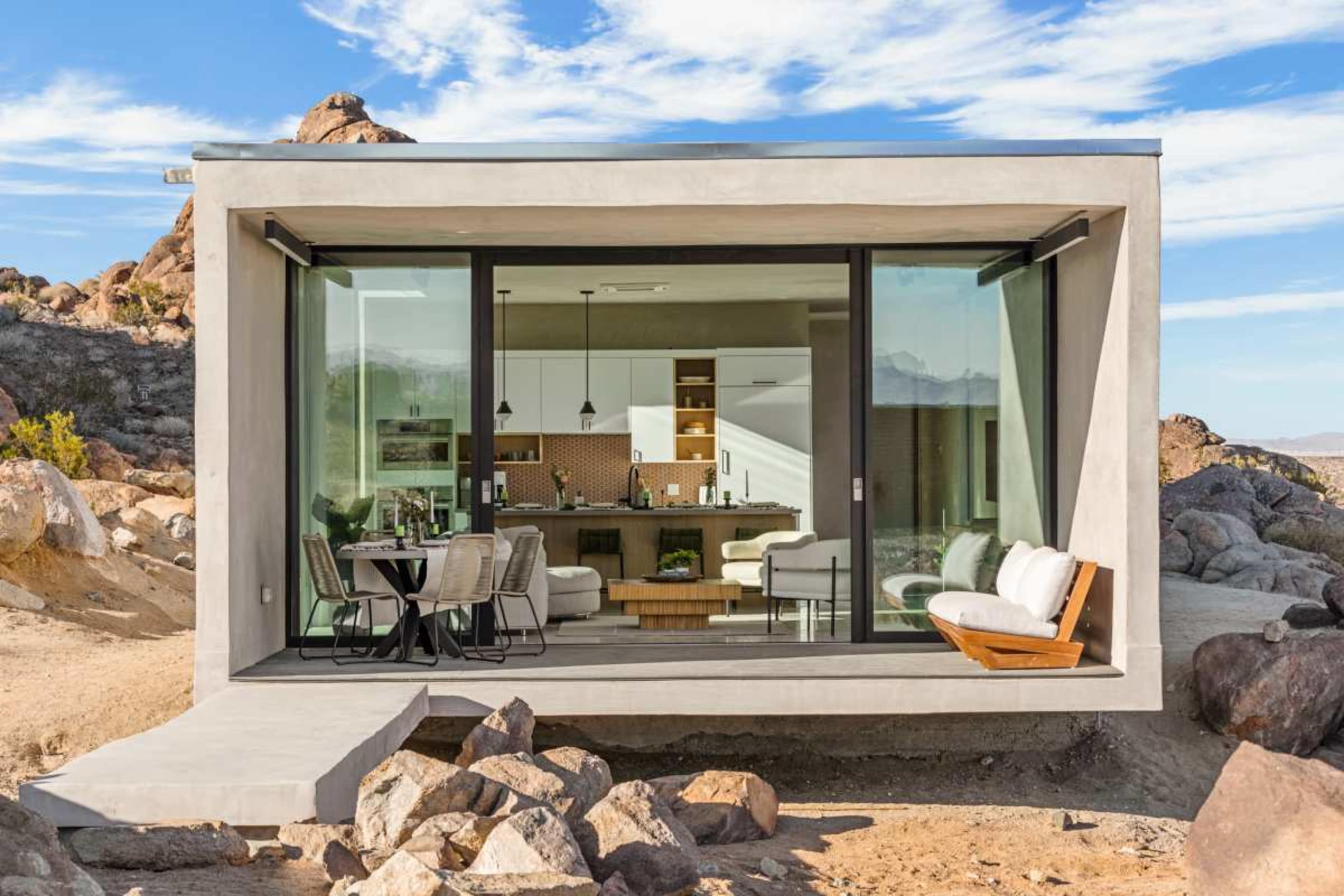 A modern, glass-fronted house sits among large rocks in a desert landscape.