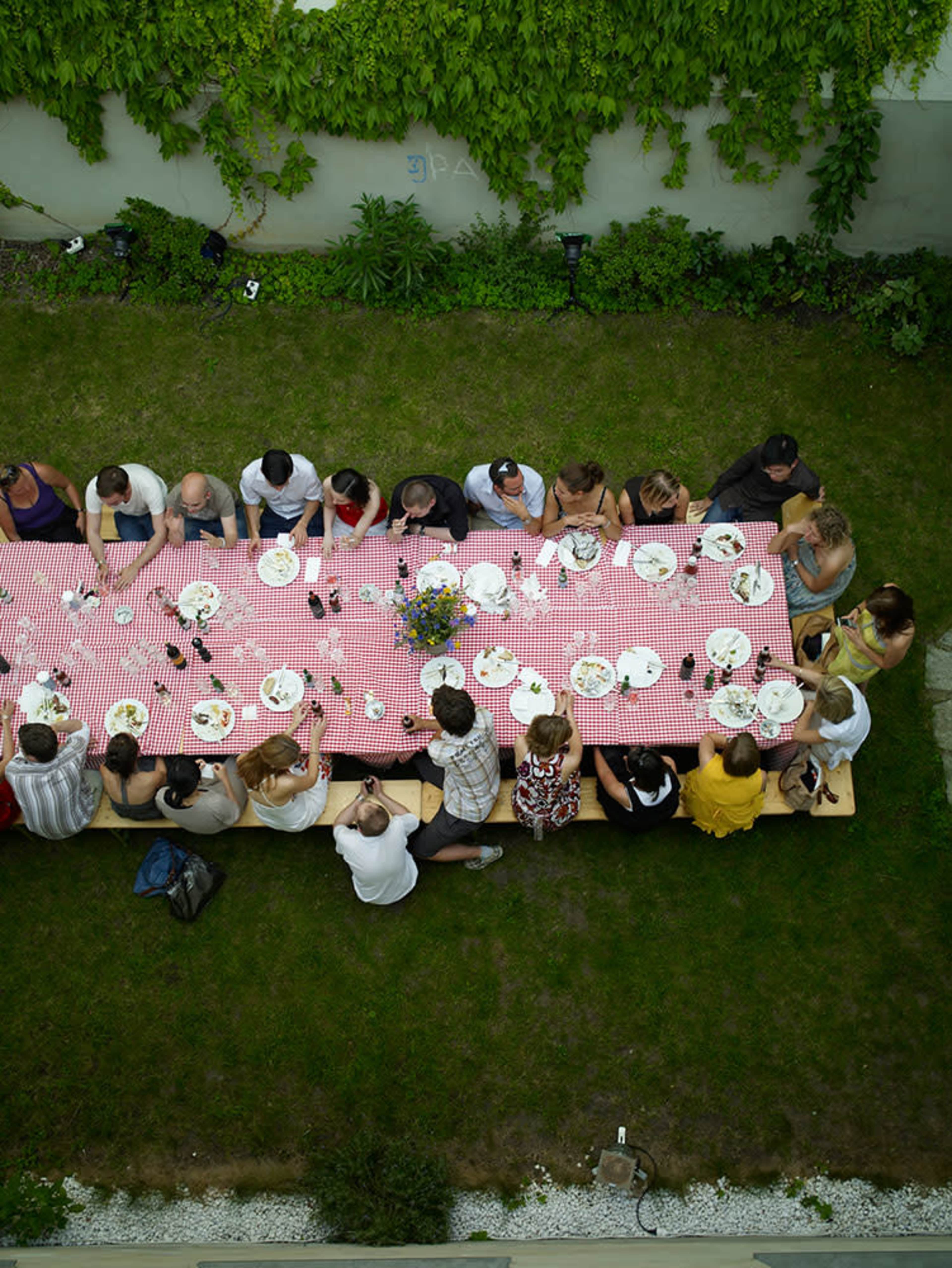 A long table covered with a red and white checkered tablecloth is filled with plates and drinks, surrounded by a group of people enjoying a meal outdoors.
