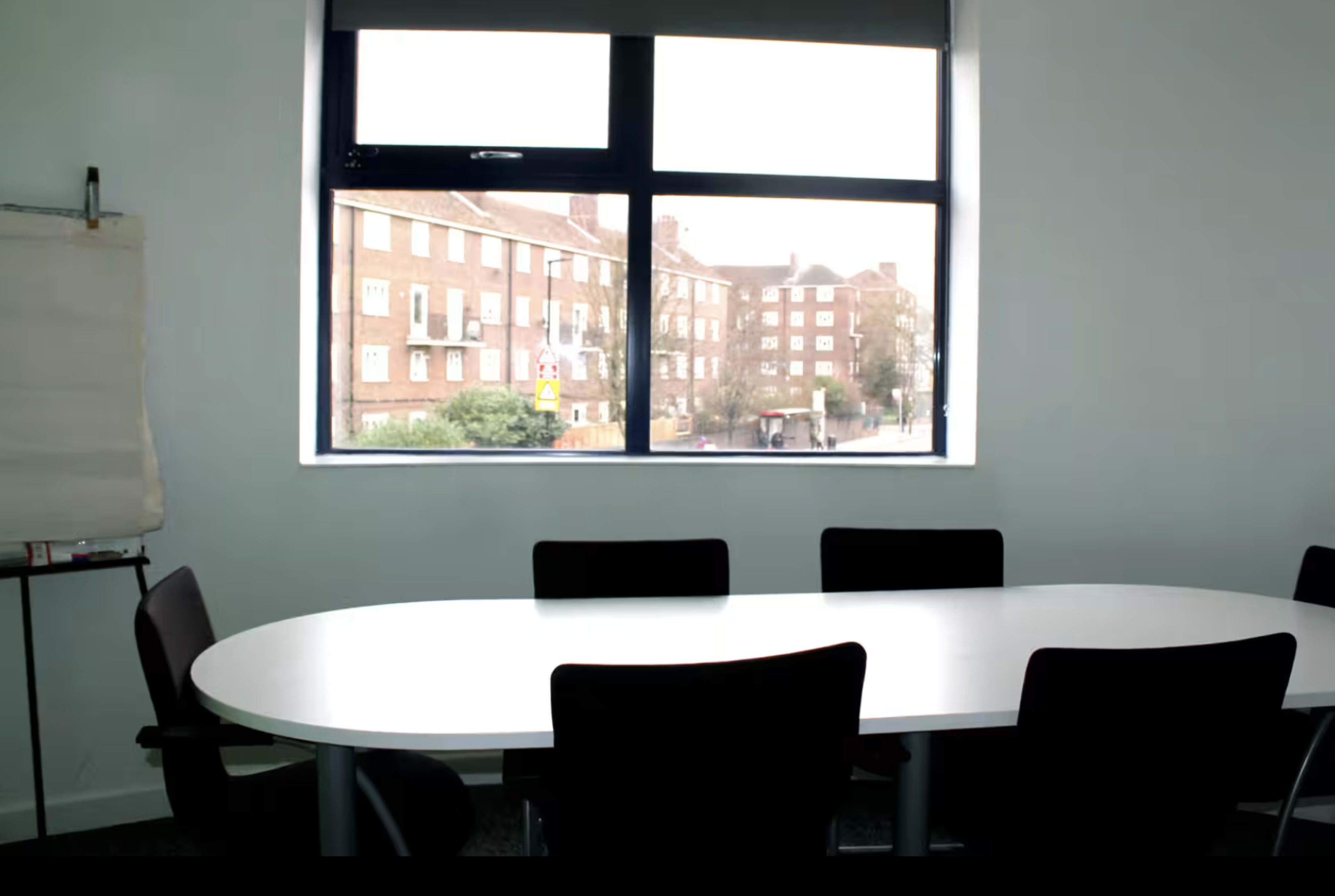 A large conference table with black chairs is positioned in a well-lit room, featuring a window that overlooks a street and several brick buildings.