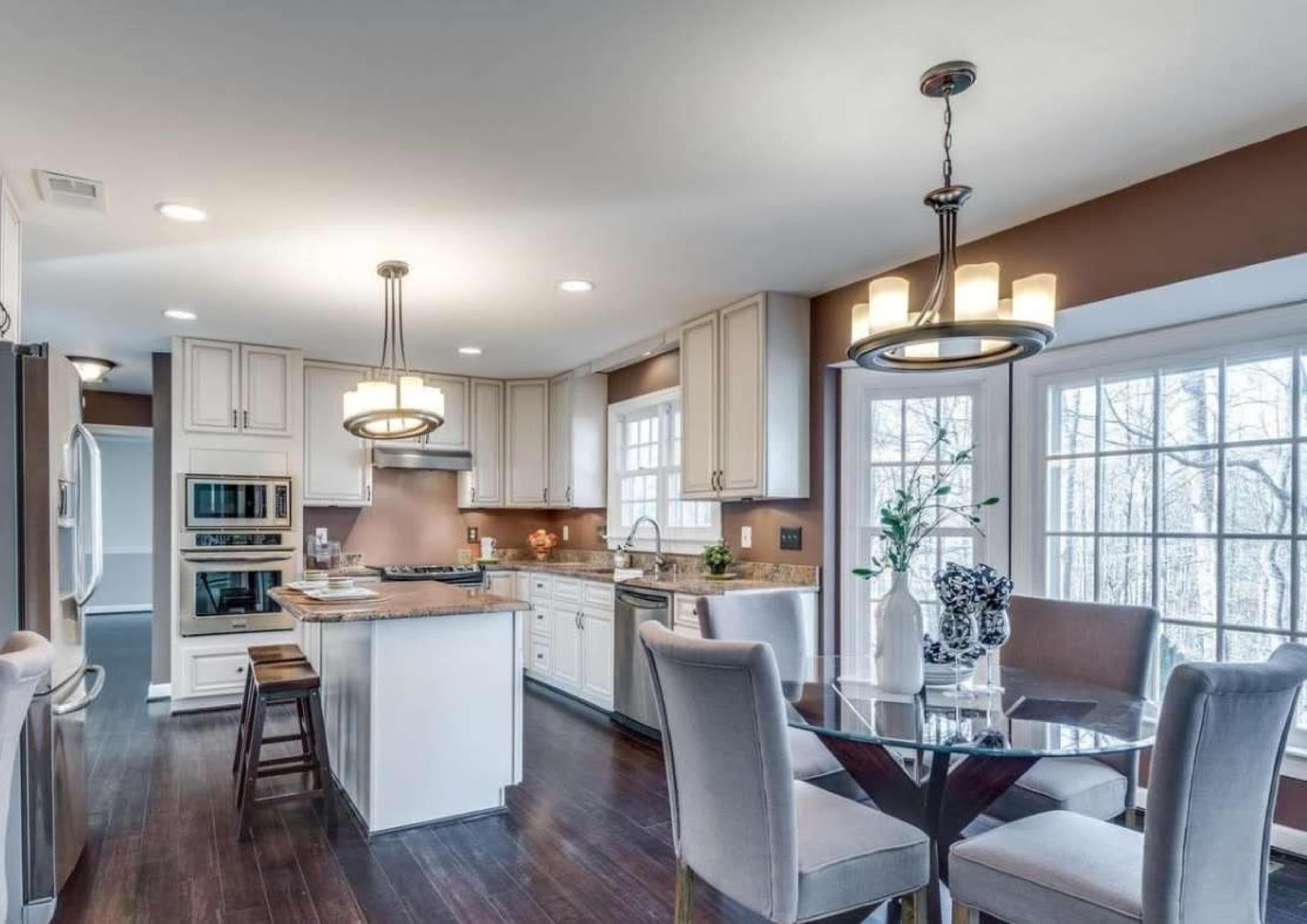 The image shows a modern kitchen with white cabinets, a central island, and a dining area featuring a round glass table and upholstered chairs.
