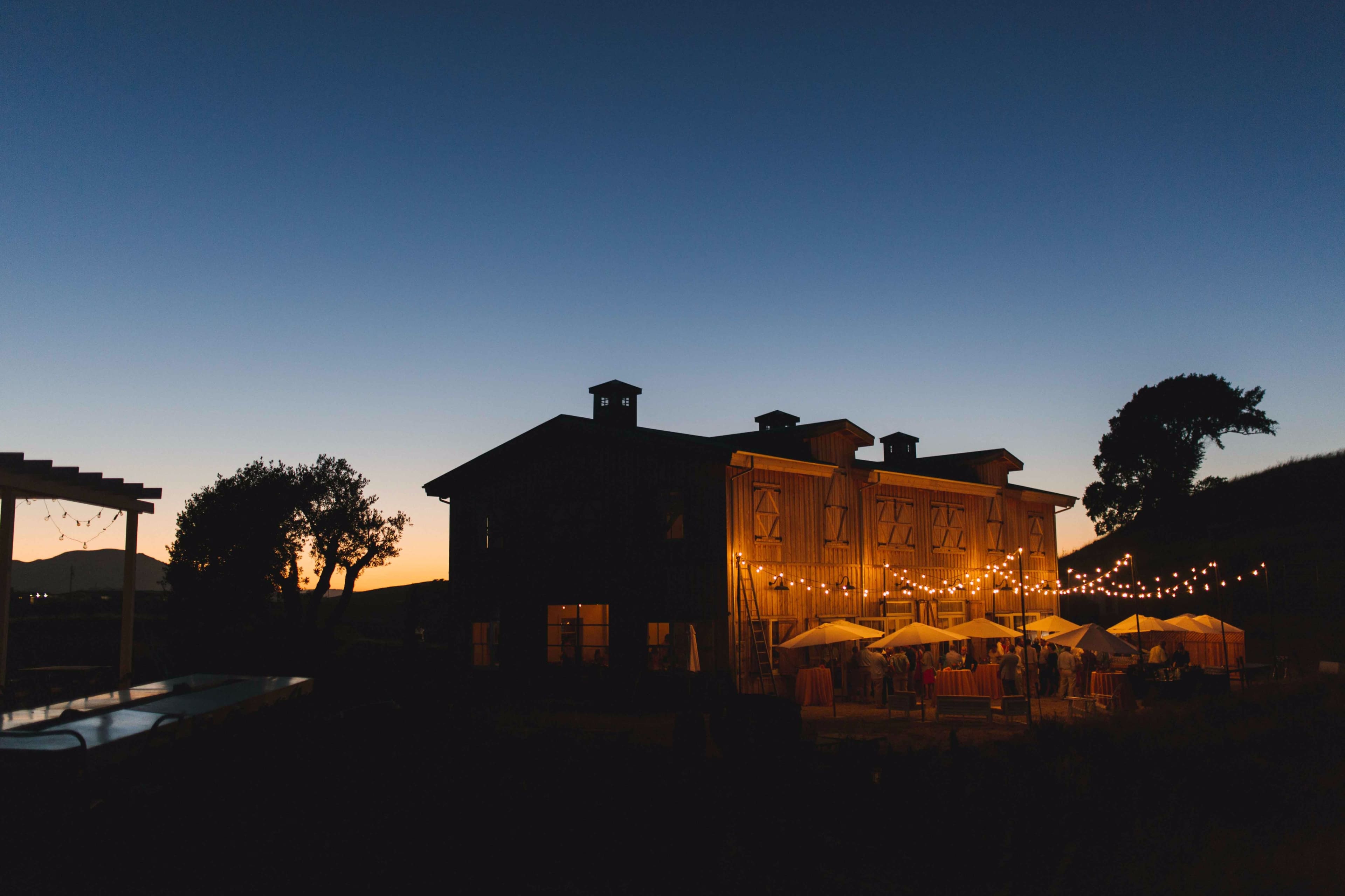 A rustic barn illuminated by string lights stands against a twilight sky with a sunset in the background.