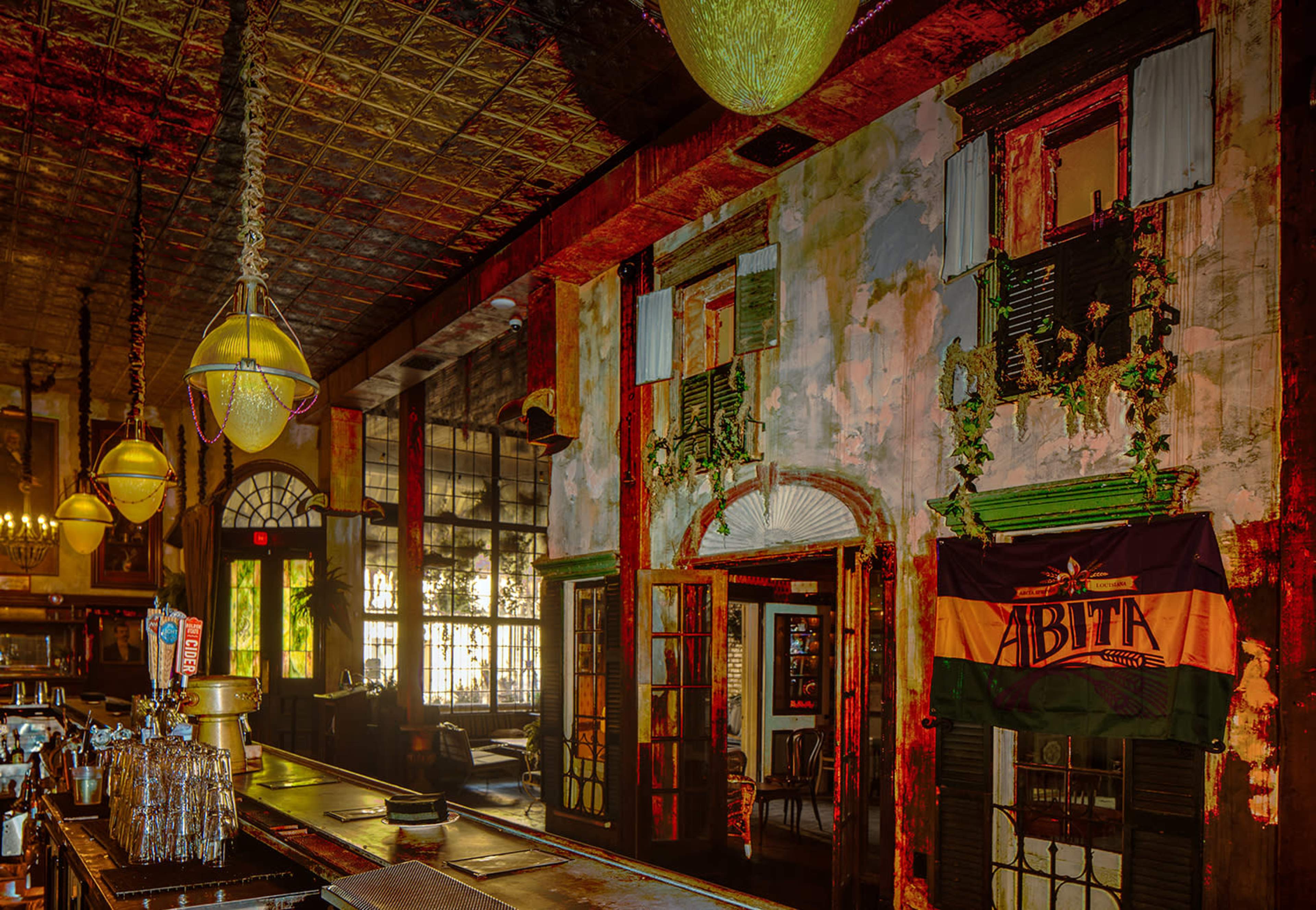 The interior of a vintage bar with exposed brick walls, large windows, and hanging pendant lights, featuring a flag and various decorative elements.