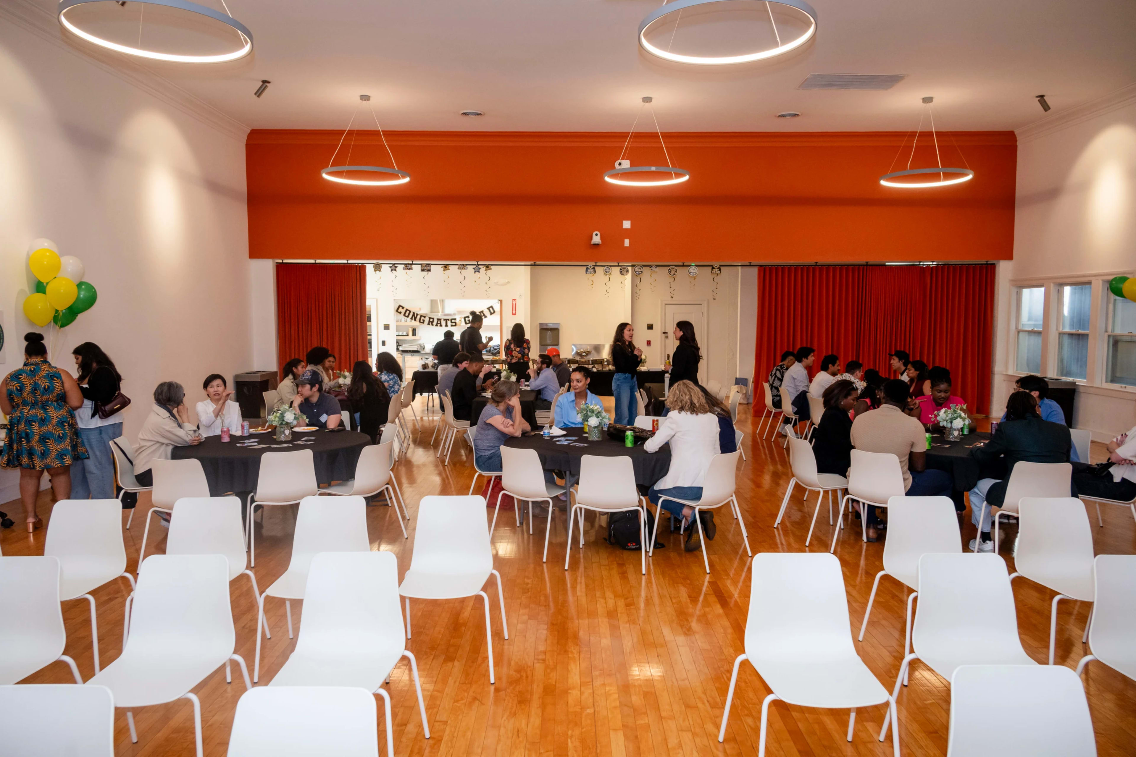 A community gathering with multiple tables set up for dining is taking place in a spacious hall decorated with balloons and banners.