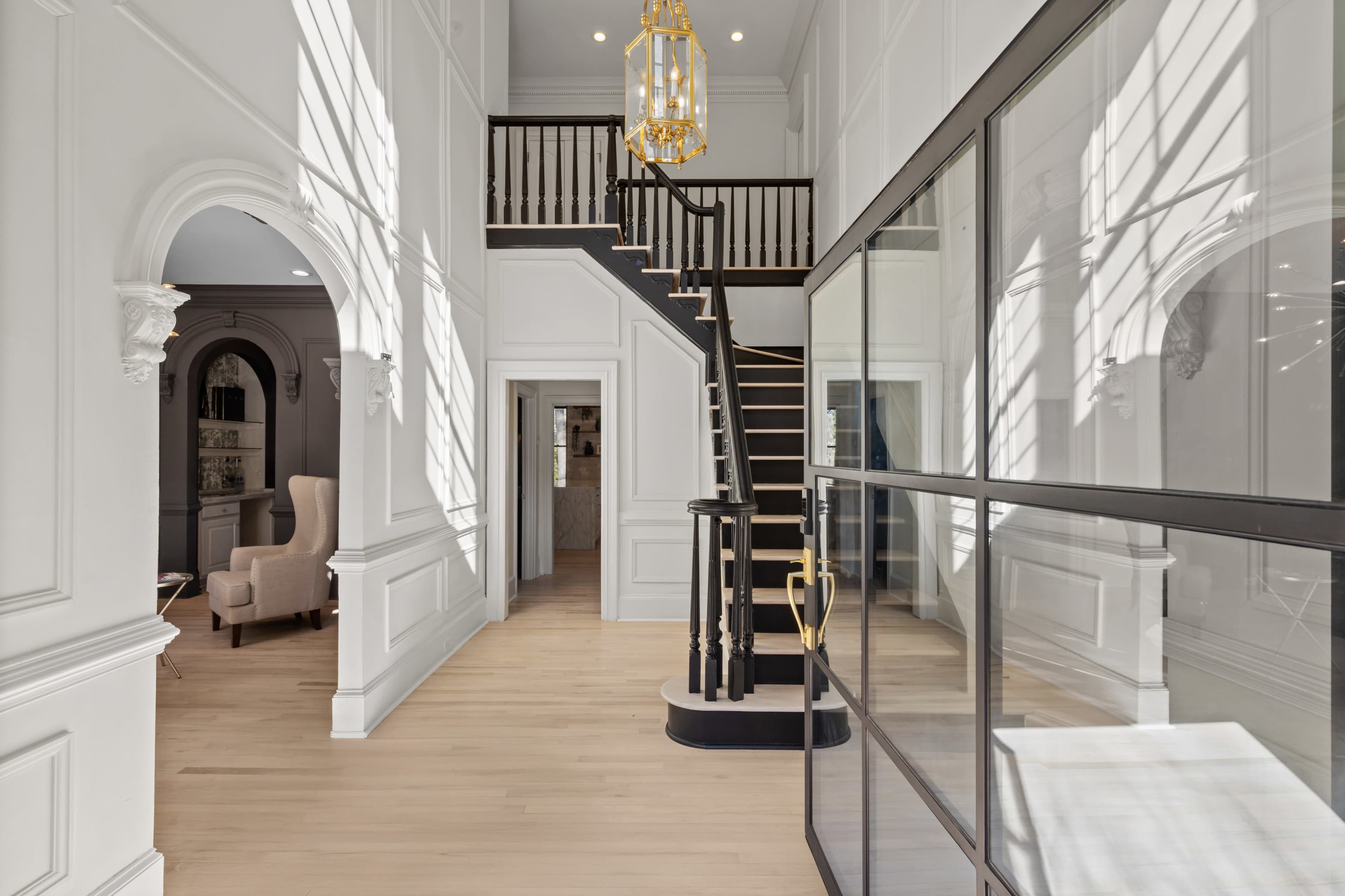 The image shows a spacious, well-lit foyer featuring a grand staircase, elegant light fixture, and large windows casting shadows on the walls.