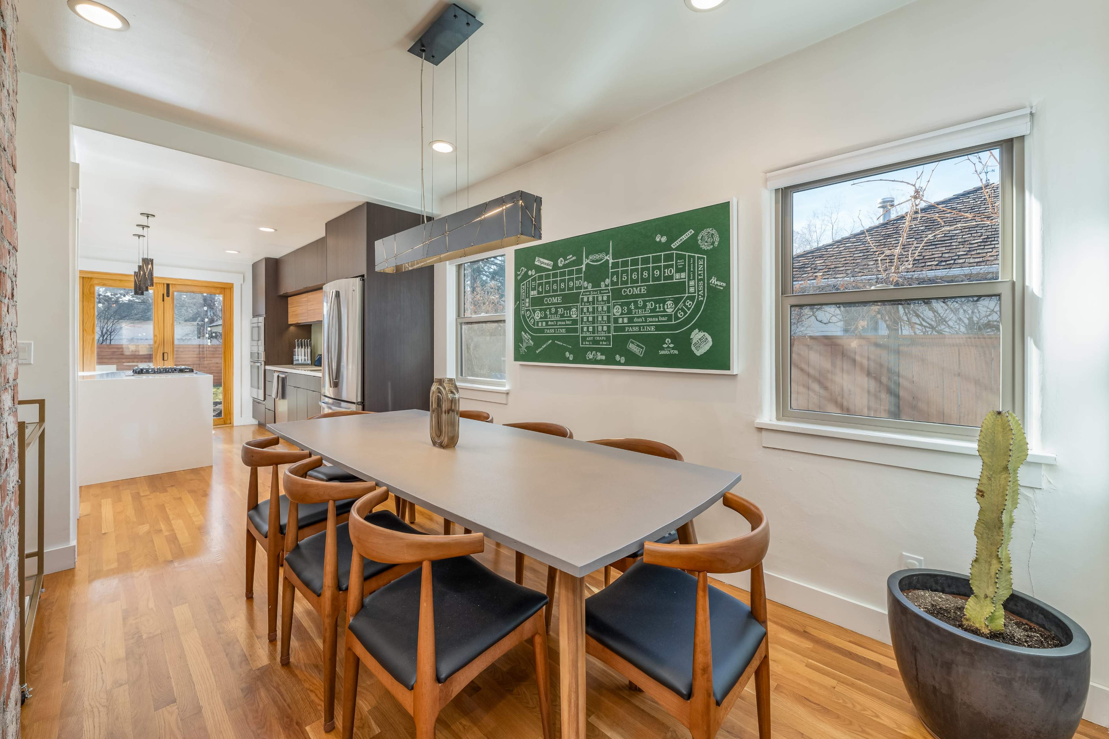 A modern dining area features a long table with wooden chairs, a chalkboard wall with a diagram, and large windows letting in natural light.