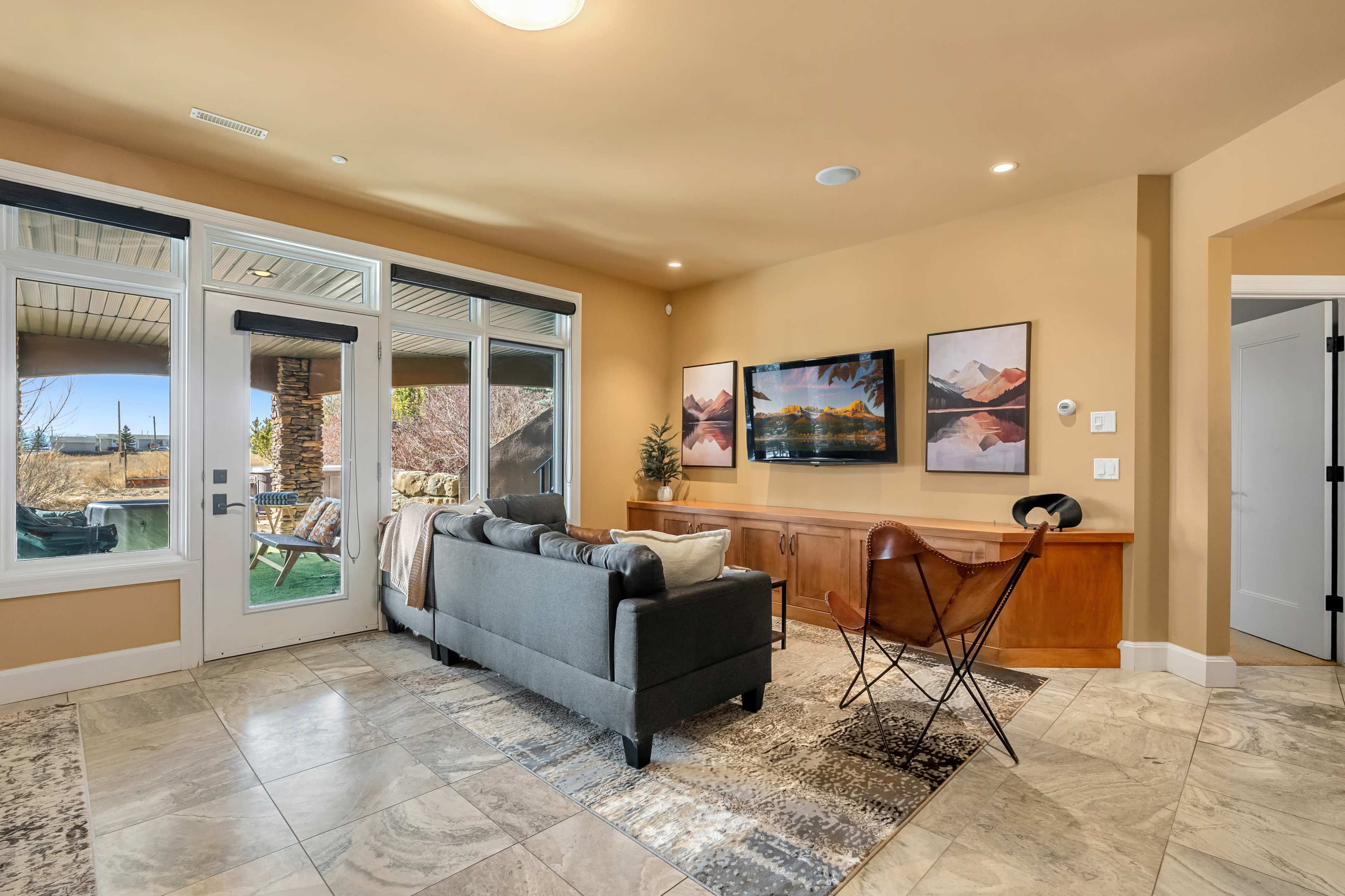 The image shows a living room with a gray sofa, a chair, a wooden sideboard, and two framed pictures on a wall, featuring large windows that provide natural light and a view outside.
