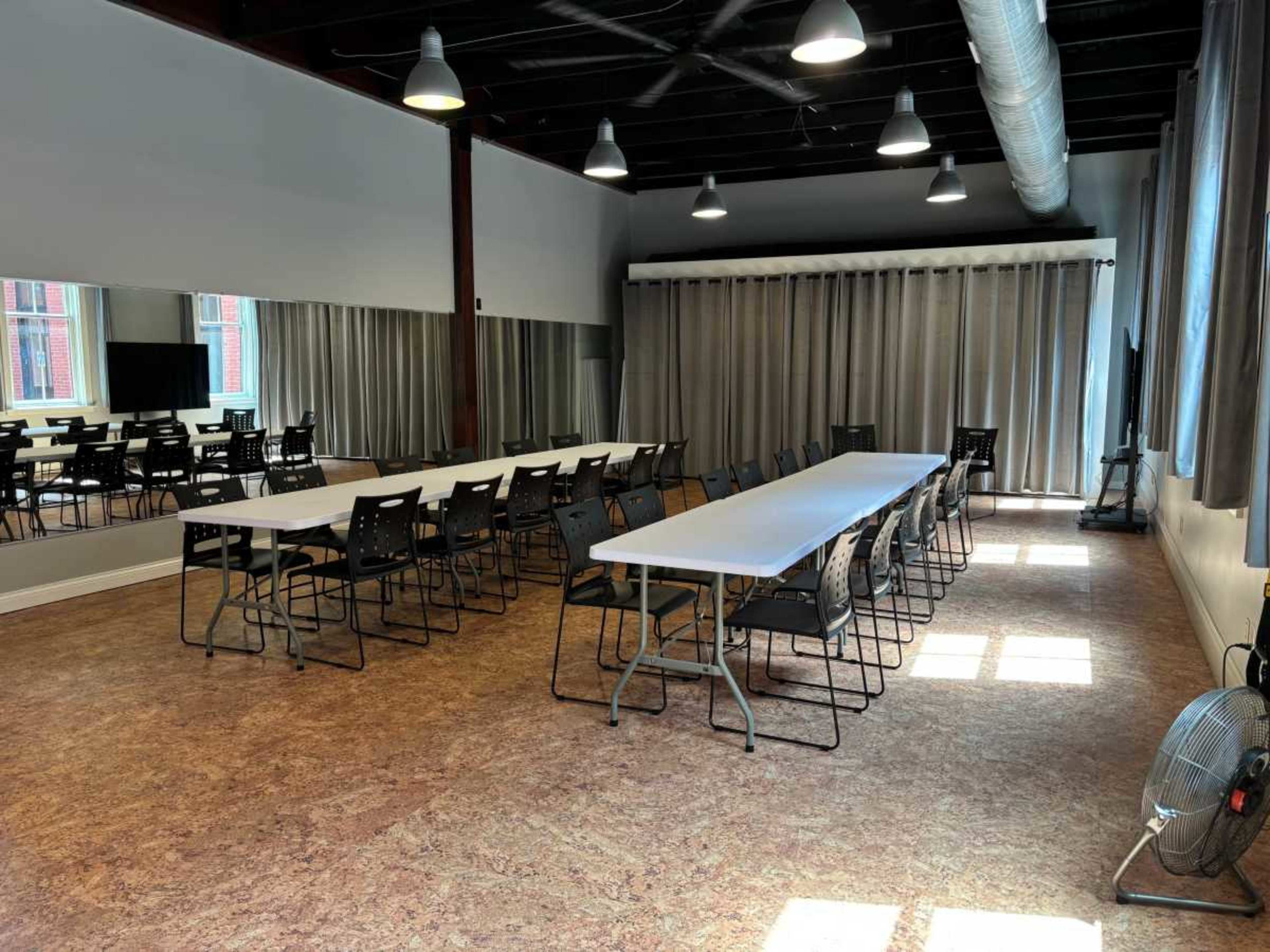 The image shows an interior meeting space with multiple tables arranged in a U-shape and chairs, large windows with curtains, and a mirror along one wall.