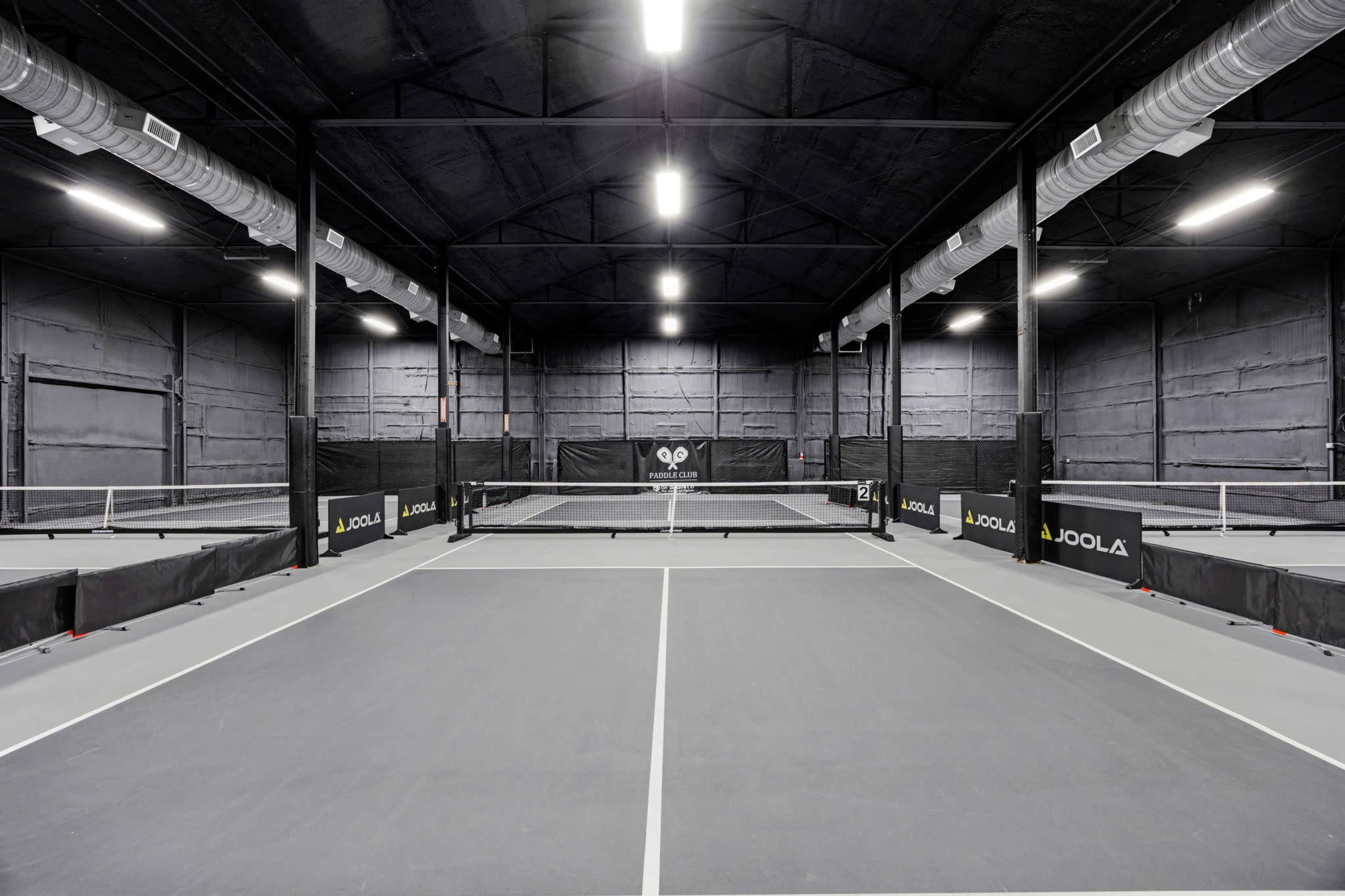The image shows a large indoor table tennis facility with multiple tables set up and a black, industrial-style interior.