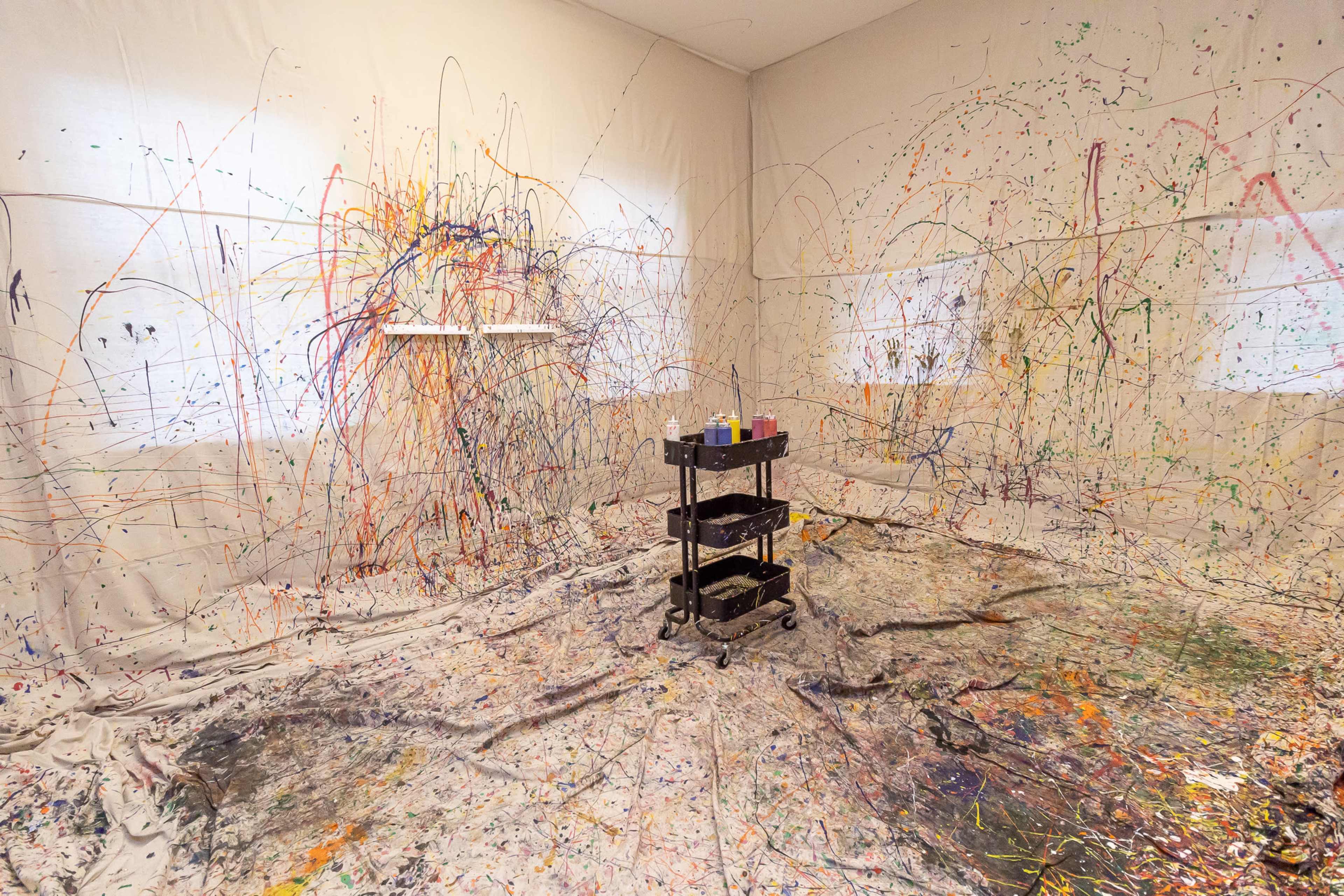 A messy art studio with splattered paint on the walls and floor, and a cart with art supplies in the center.