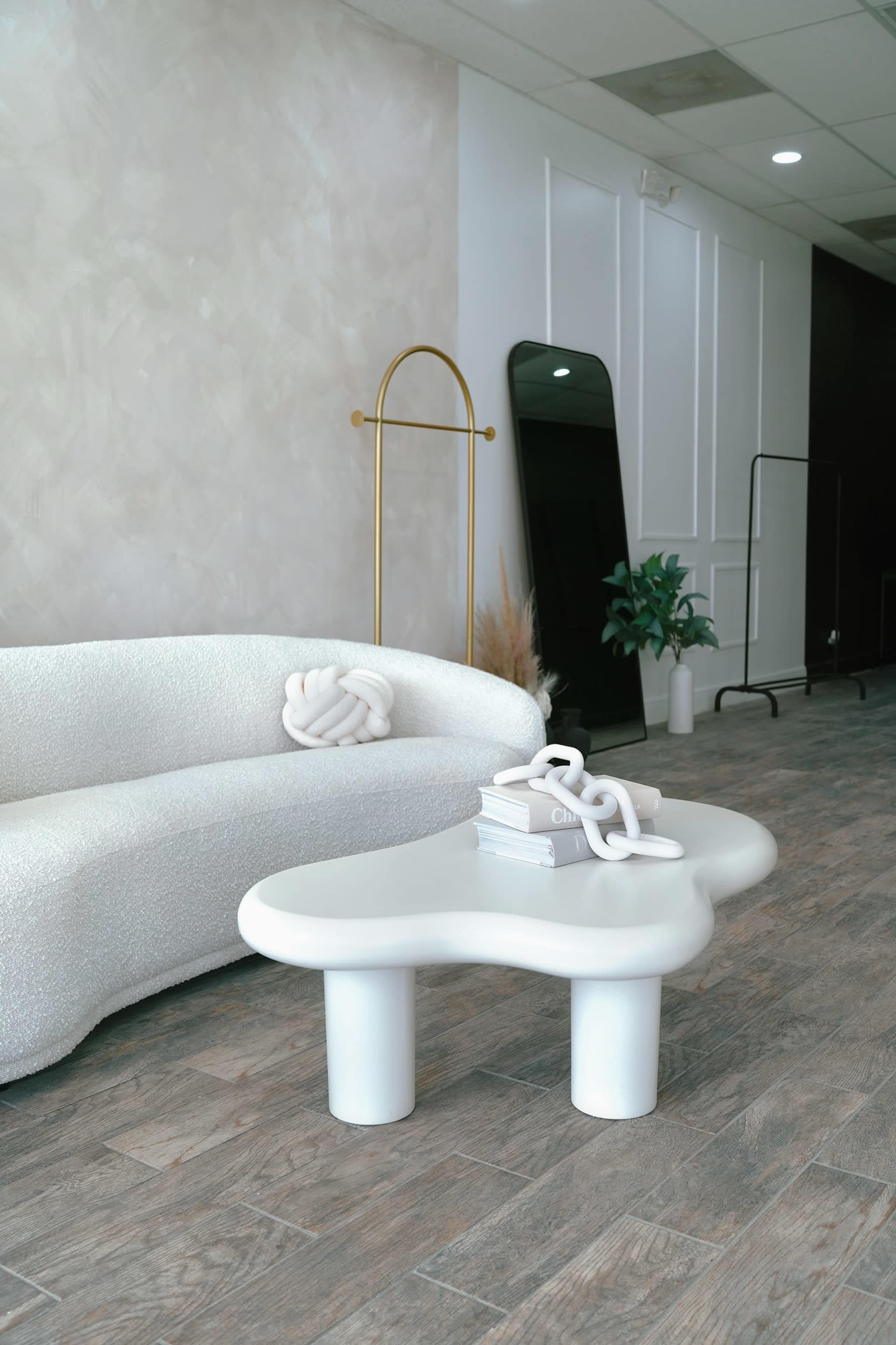 A modern living space features a white, sculptural sofa and an organic-shaped white coffee table, accompanied by a large mirror and a potted plant.