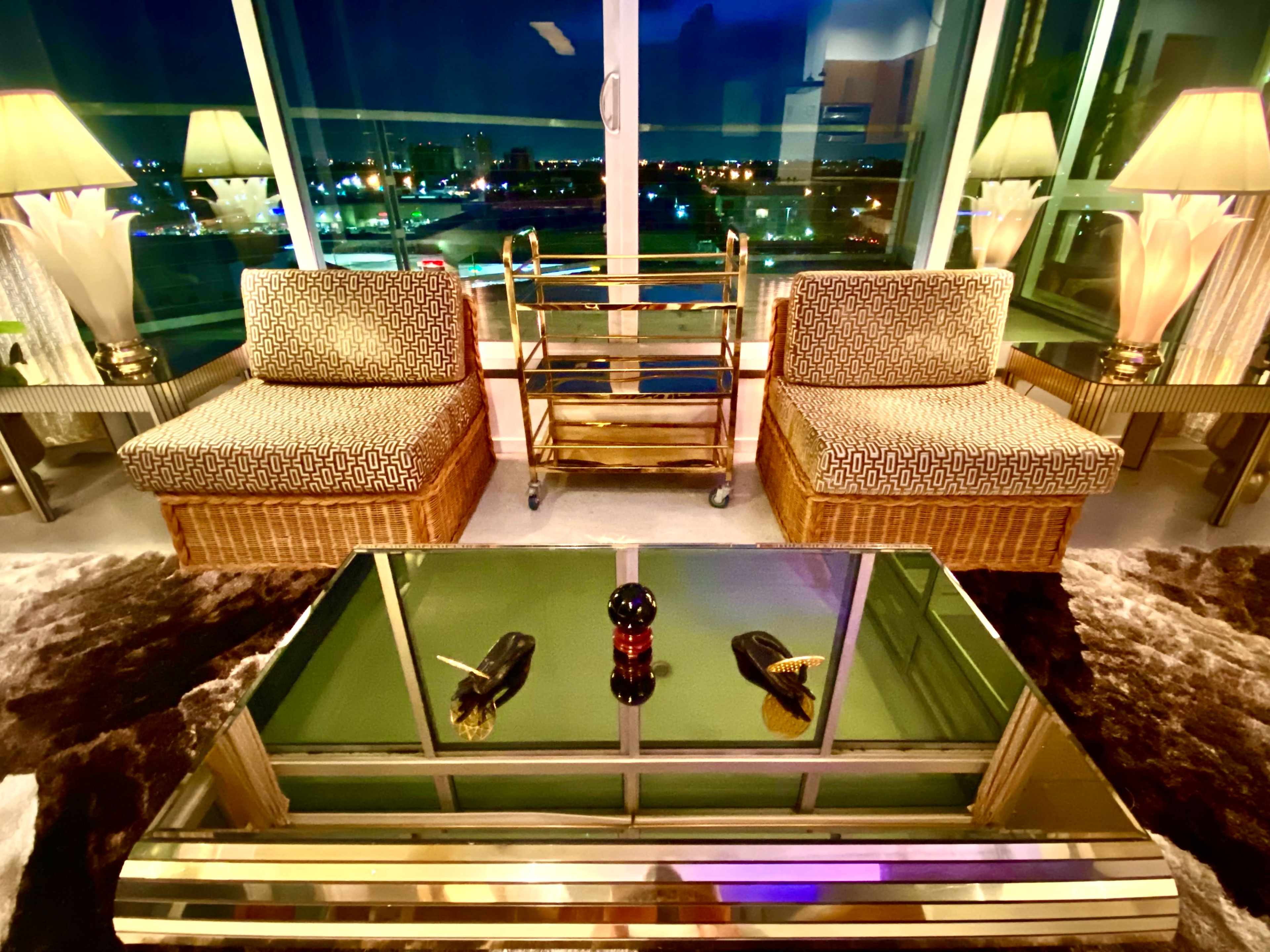 The image shows a stylish living room featuring two patterned chairs, a glass coffee table with a decorative item, and a wheeled cart, all set against a backdrop of city lights at night.