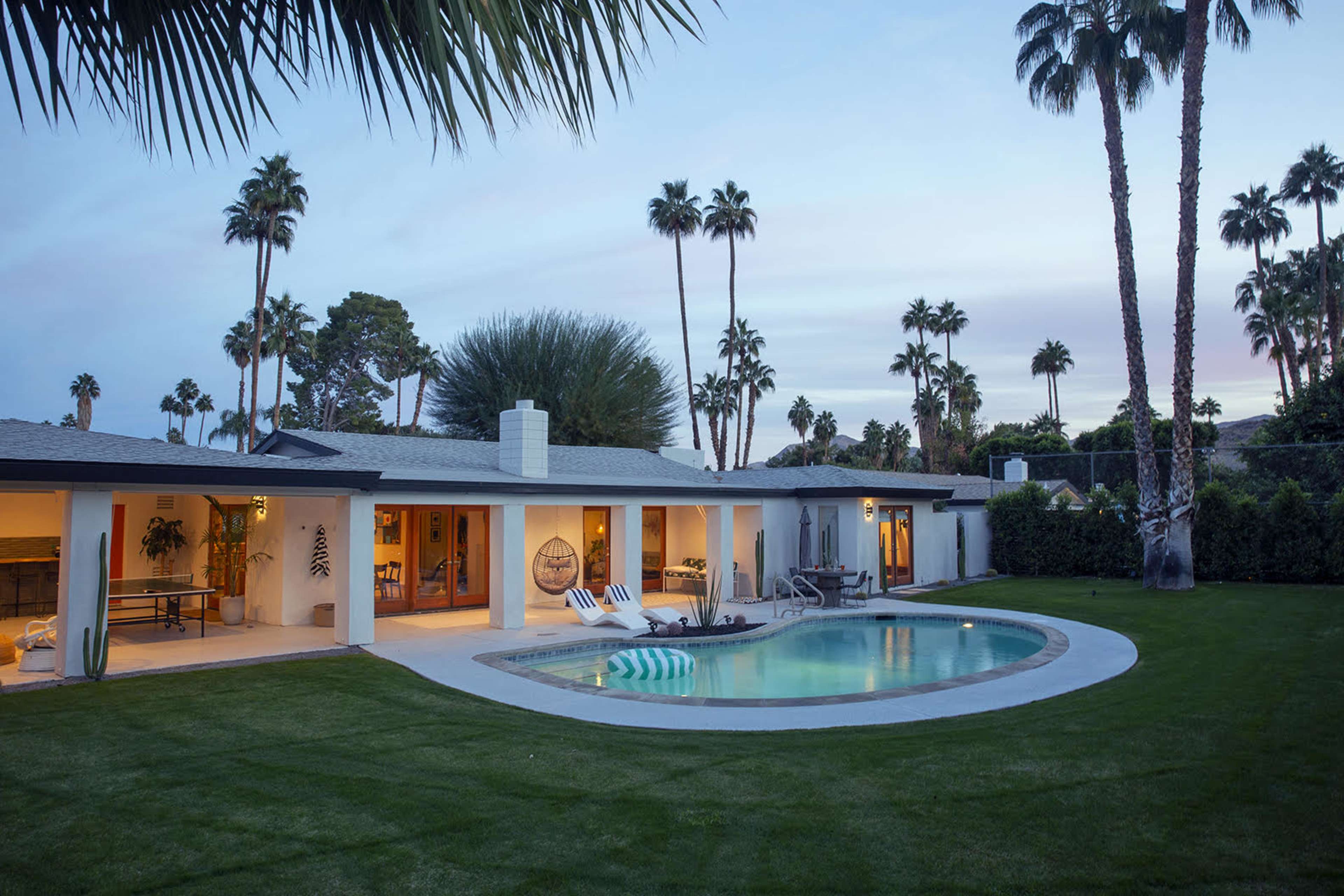 A modern house with a pool is surrounded by palm trees and lush green grass during twilight.