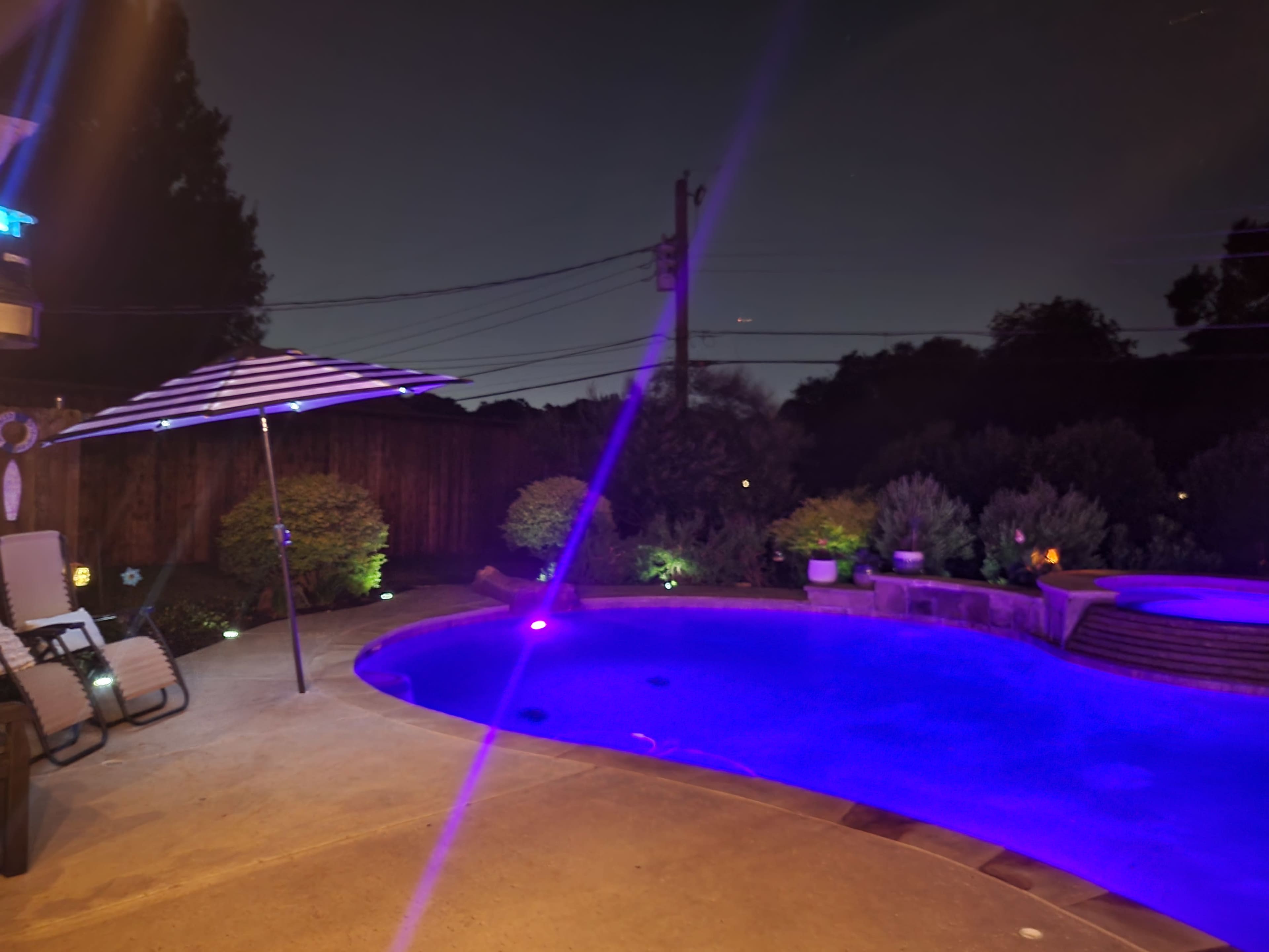 A pool with purple lighting is surrounded by landscaped greenery and features a shaded seating area under a striped umbrella at dusk.