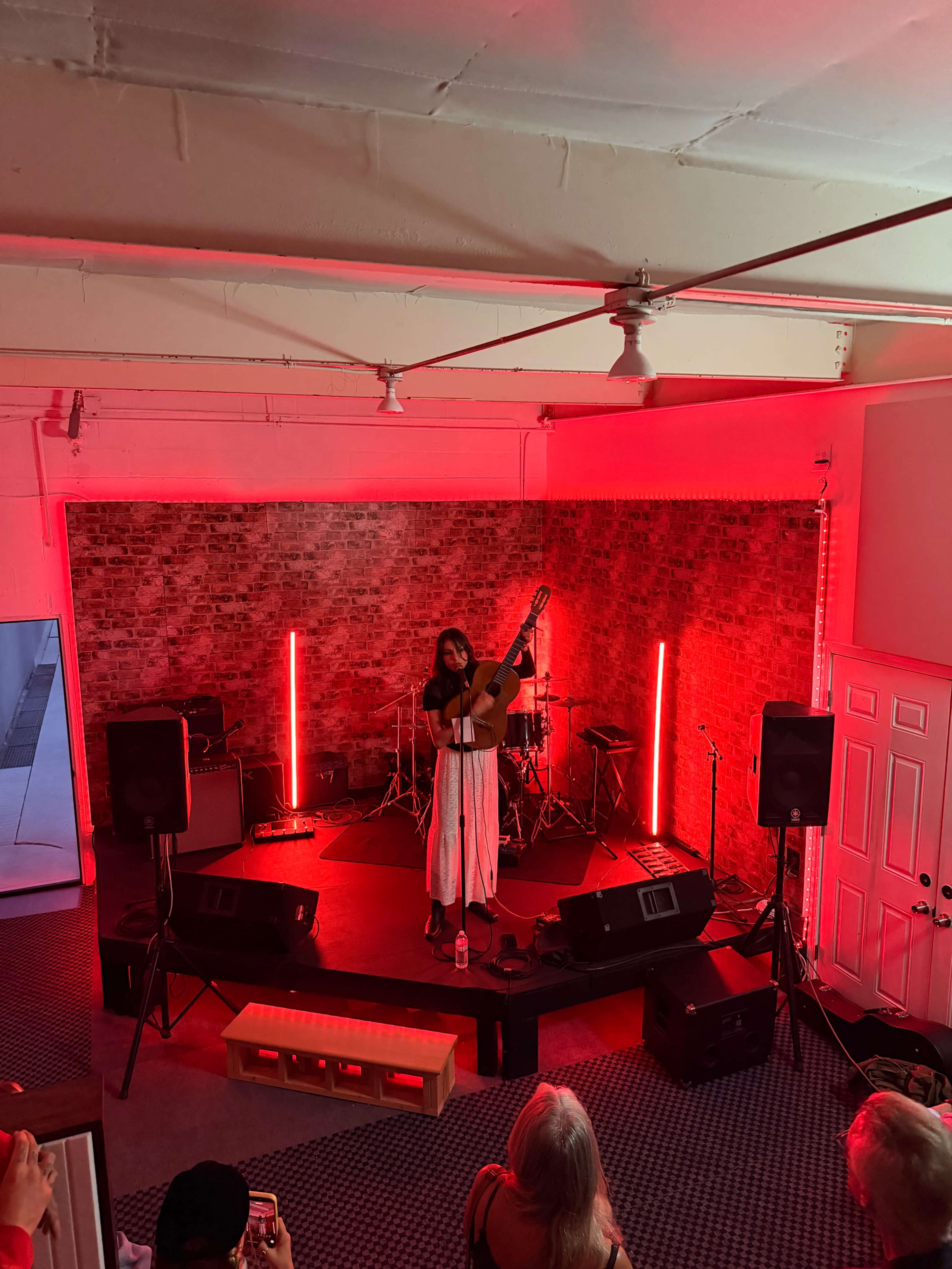 A solo musician performs with a guitar on a small stage surrounded by red lighting and a drum set in the background.