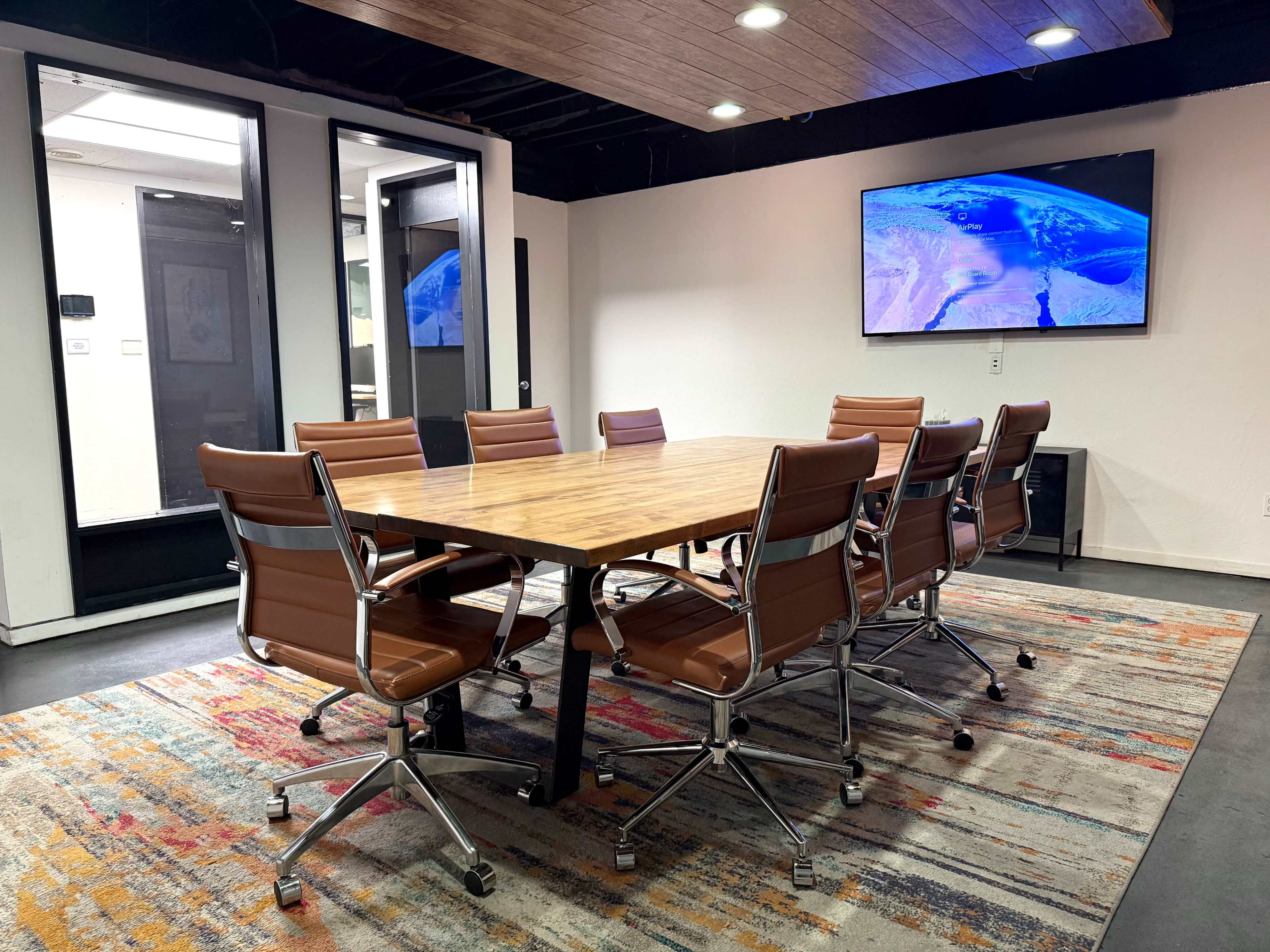 A conference room features a wooden table surrounded by eight leather chairs, with a large wall-mounted screen displaying a map in the background.