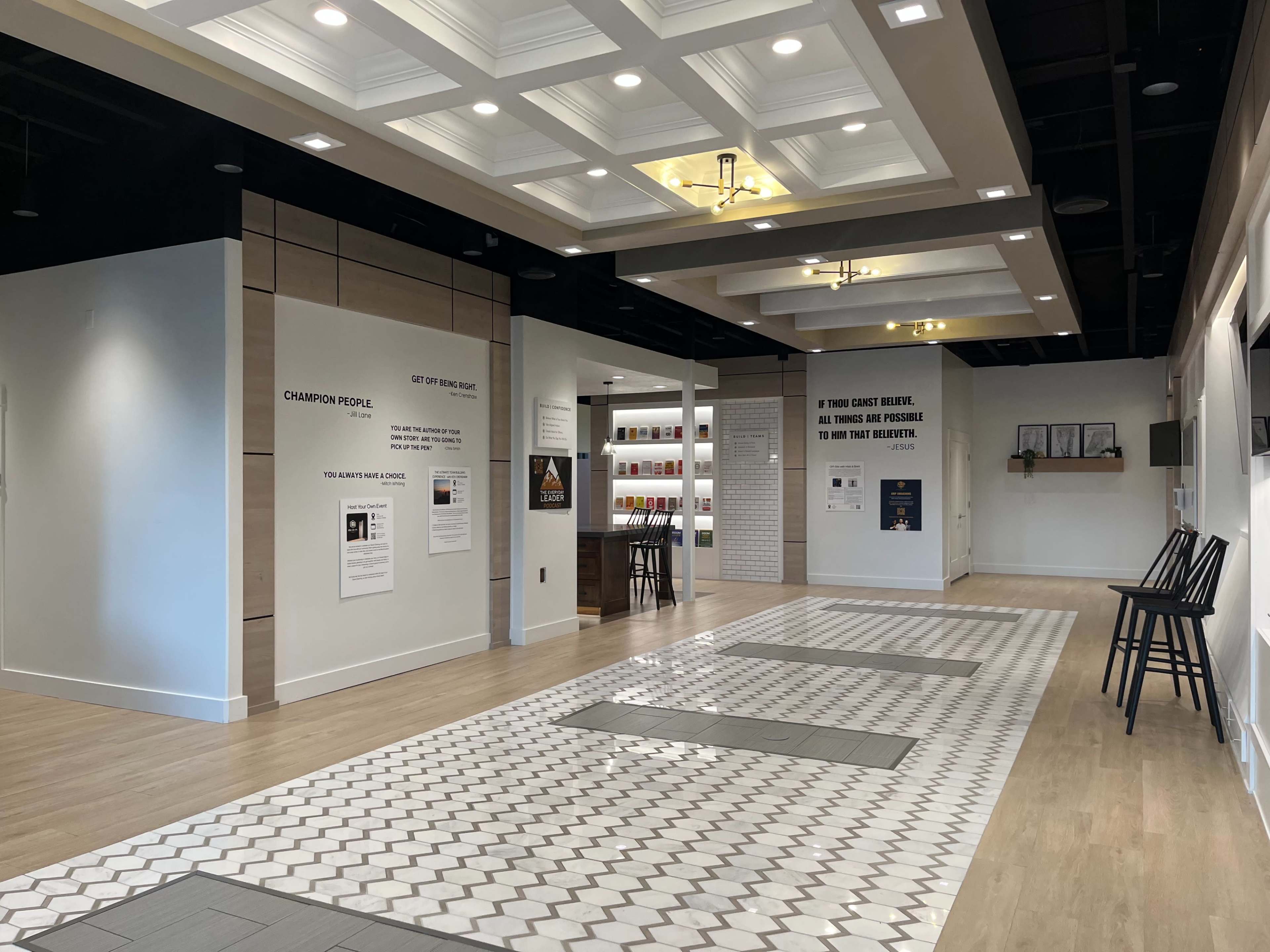 The image shows a modern, well-lit interior space featuring a tiled floor, white walls, and a seating area with chairs, along with informational displays on the walls.