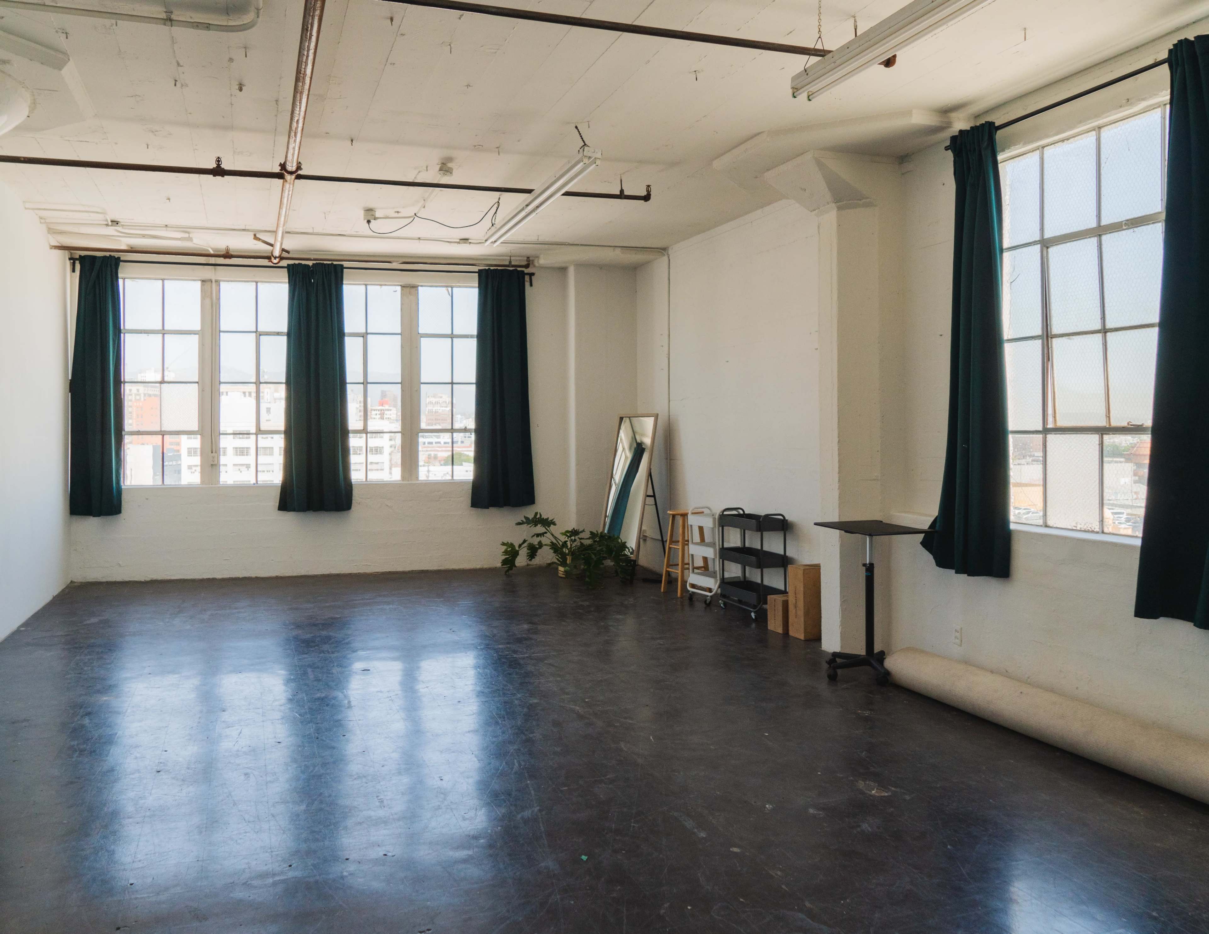 The image shows a spacious, sunlit room with large windows, green curtains, a mirror, a small table, and a roll of material on the floor.