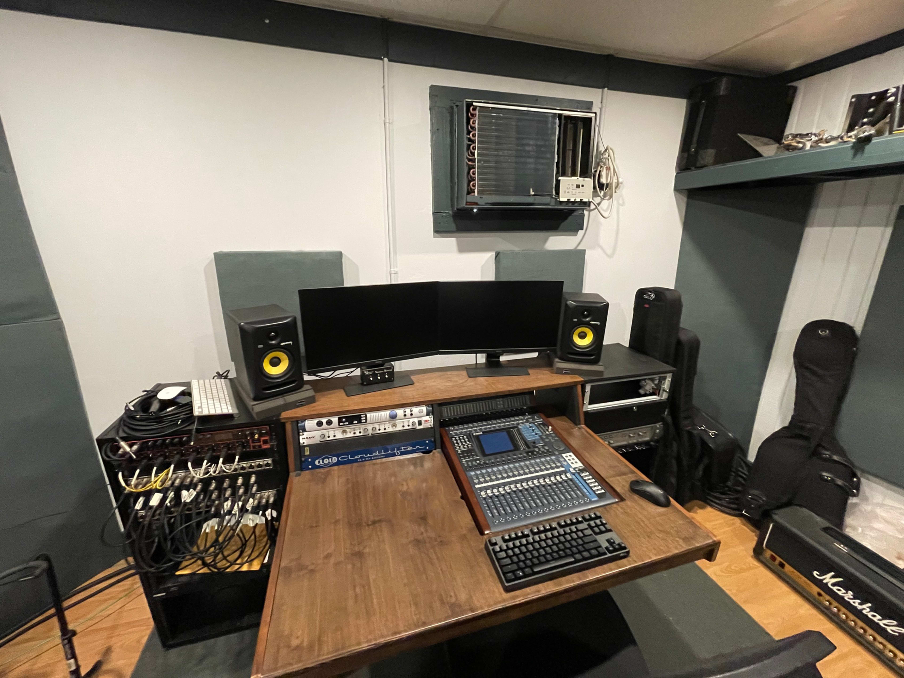 A recording studio featuring a large mixing console, two computer monitors, audio equipment, and various music instruments stored in cases.