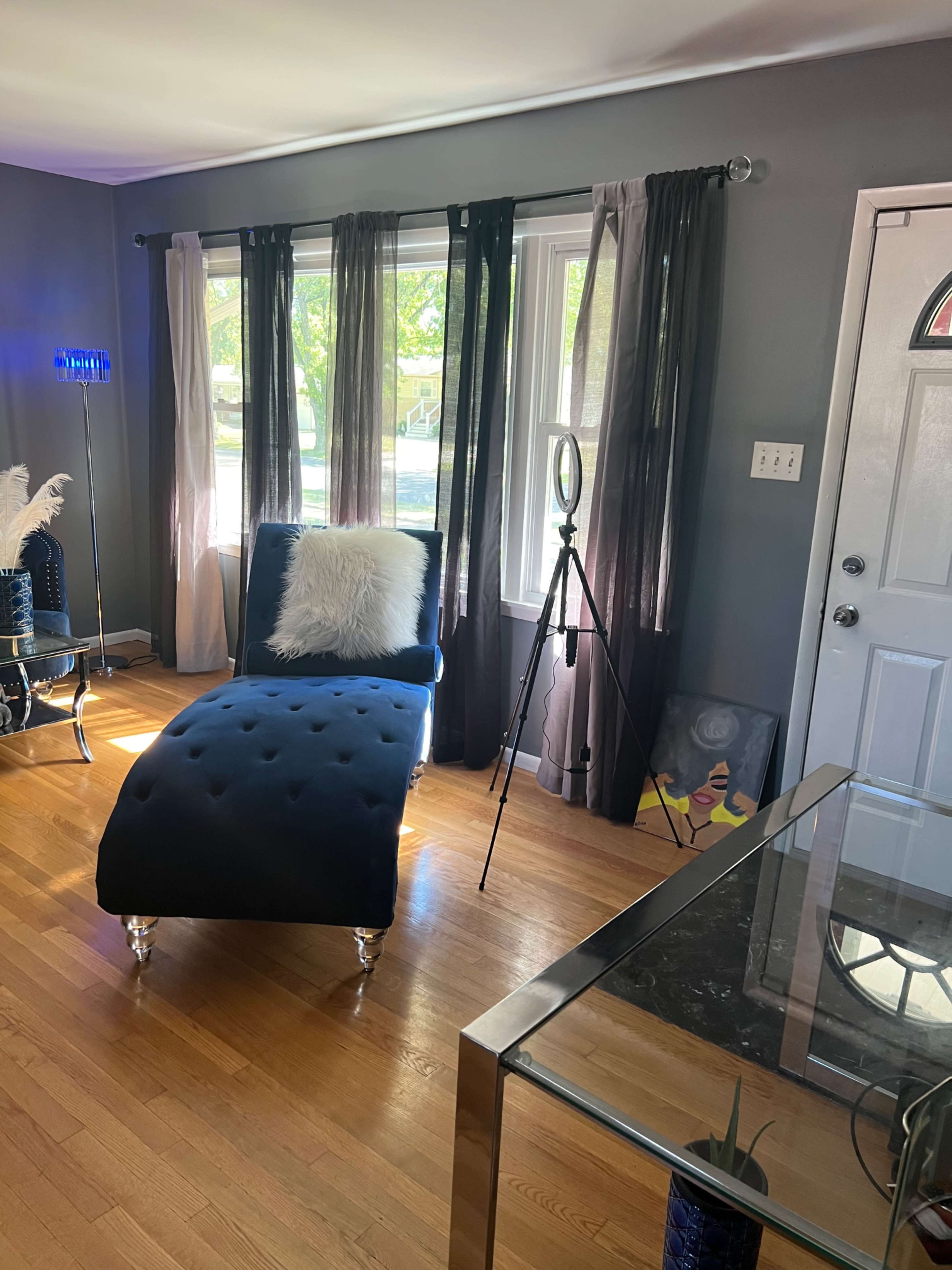 A blue chaise lounge with a white throw pillow is positioned beside a window in a room featuring gray walls and light wood flooring.