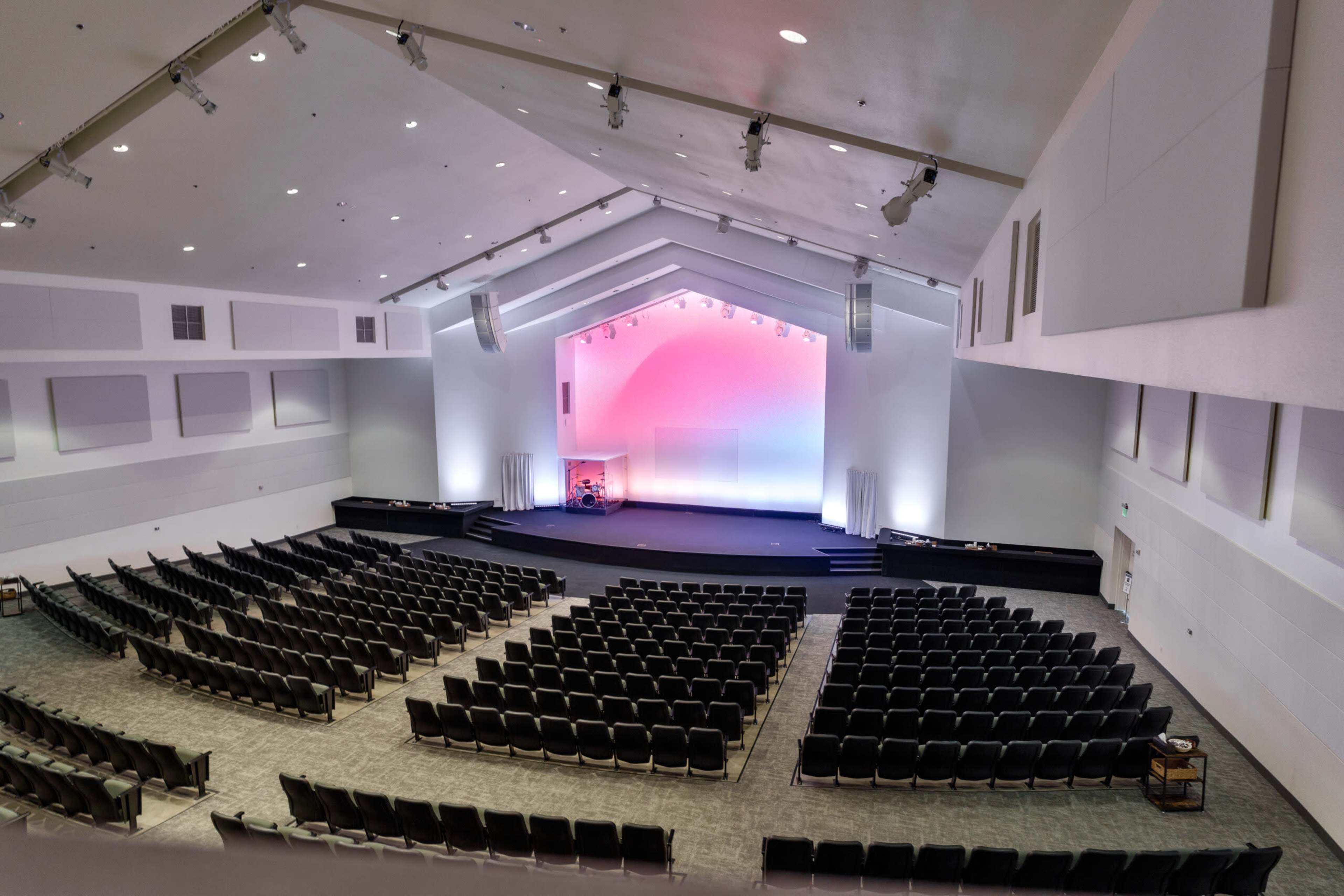The image shows a spacious auditorium with tiered seating and a stage at the front, illuminated by soft colored lights.