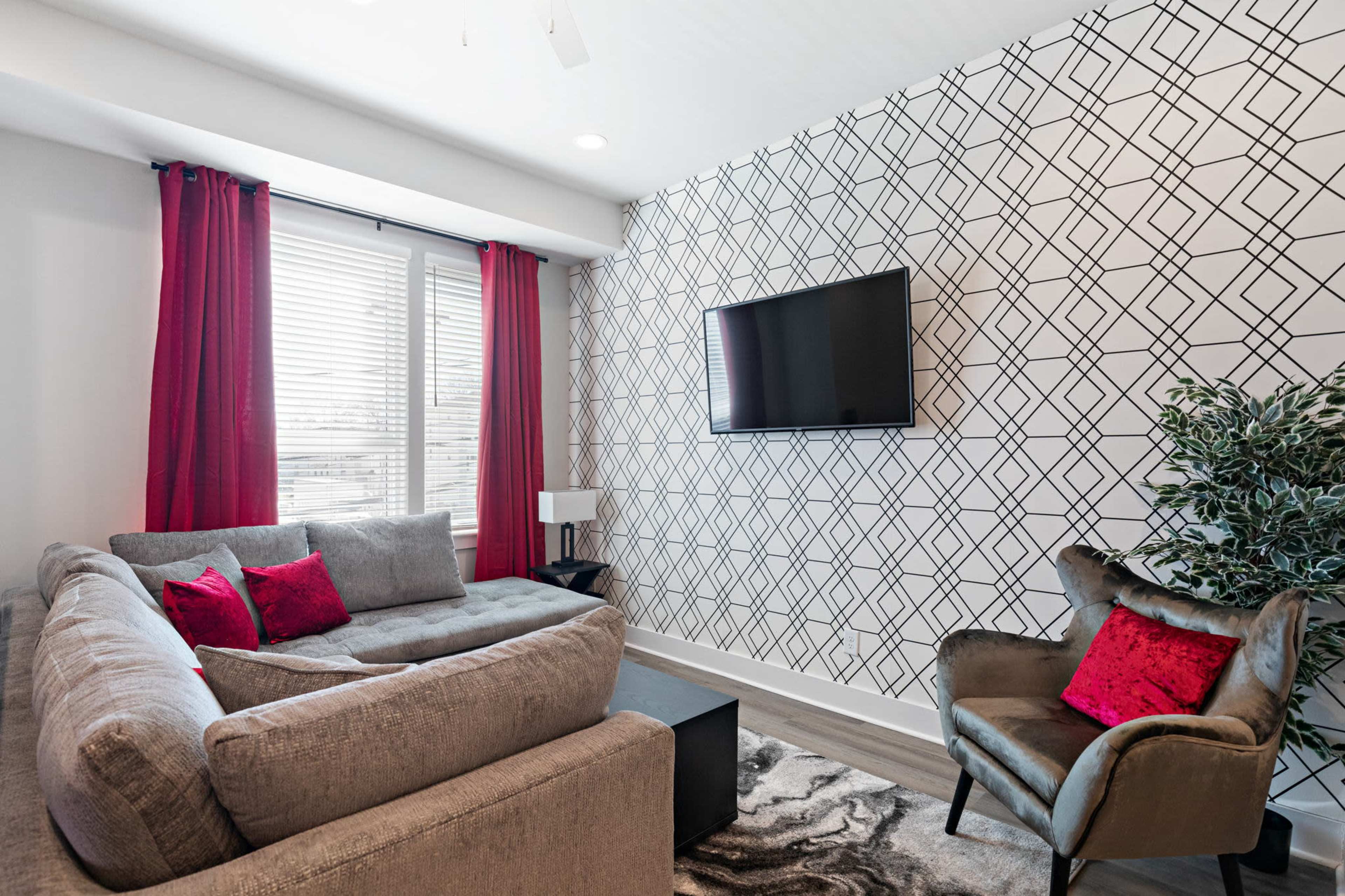 A modern living room features a gray sectional sofa with red accent pillows, a black coffee table, a flat-screen TV mounted on a patterned wall, and a decorative indoor plant.
