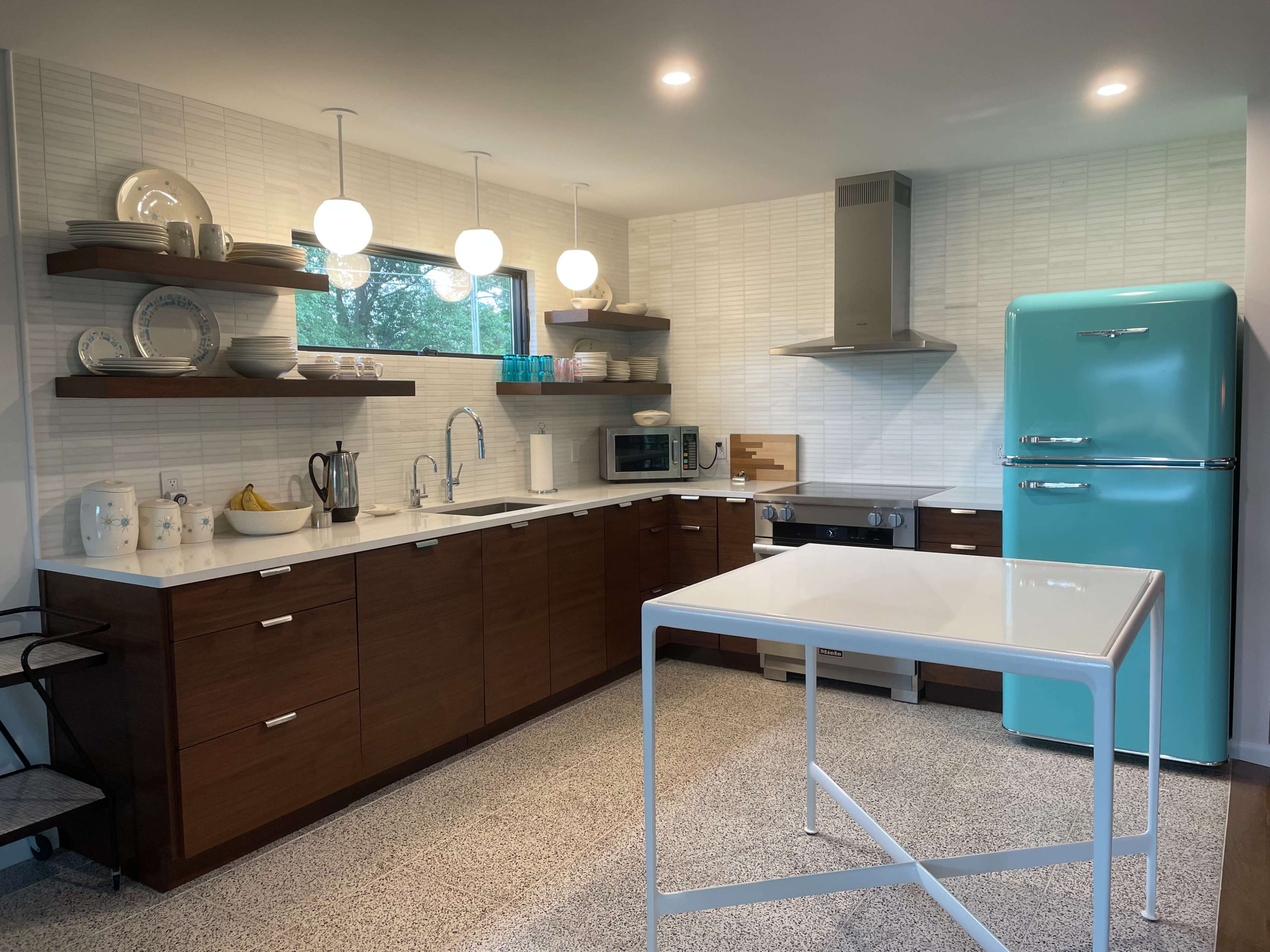 The kitchen features modern cabinets, a light blue refrigerator, and a central white table, with pendant lighting above and open shelving displaying dishware.