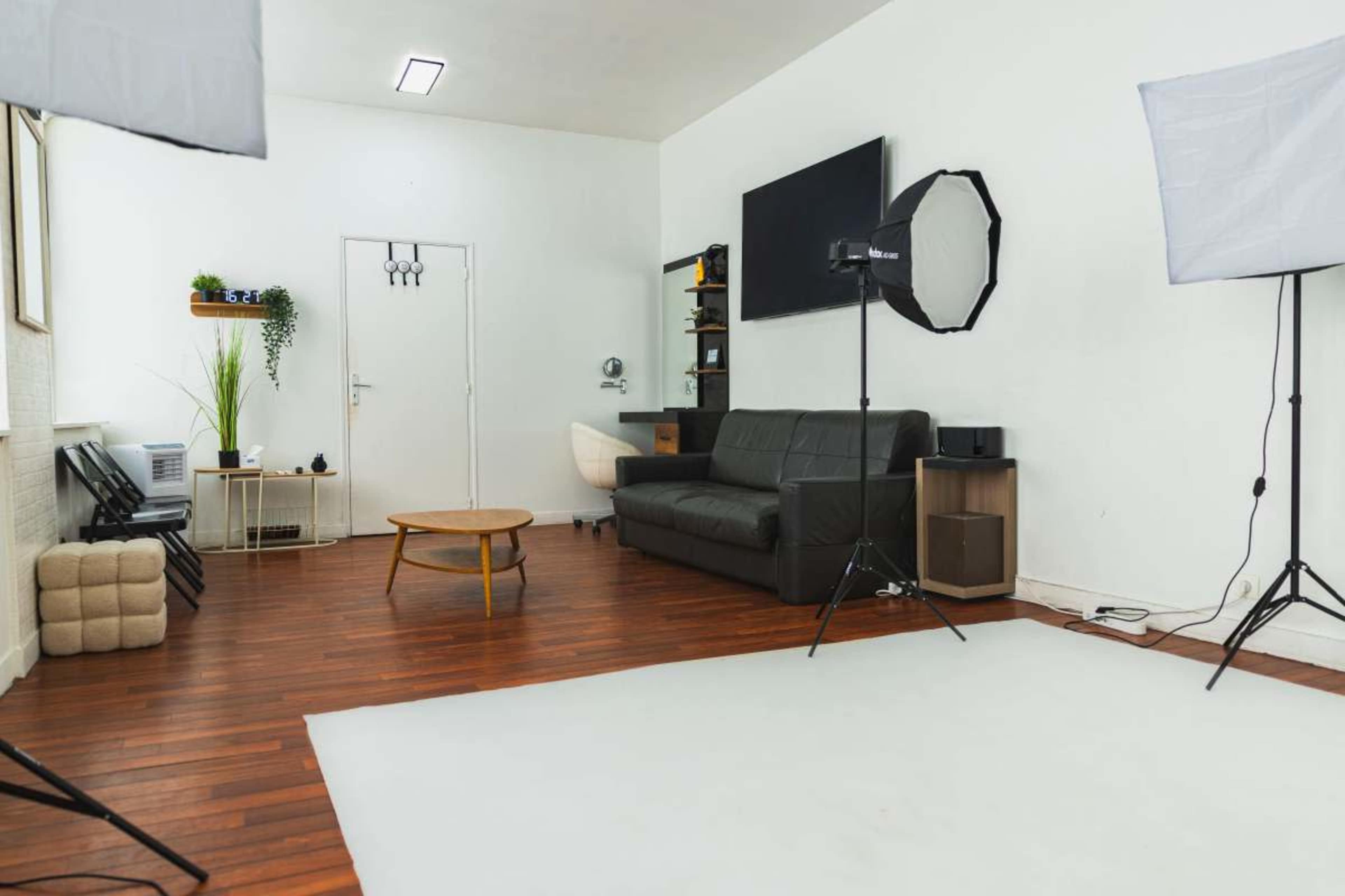 A spacious and well-lit photo studio features a black leather sofa, a wooden coffee table, and two softbox lights set up on tripods.