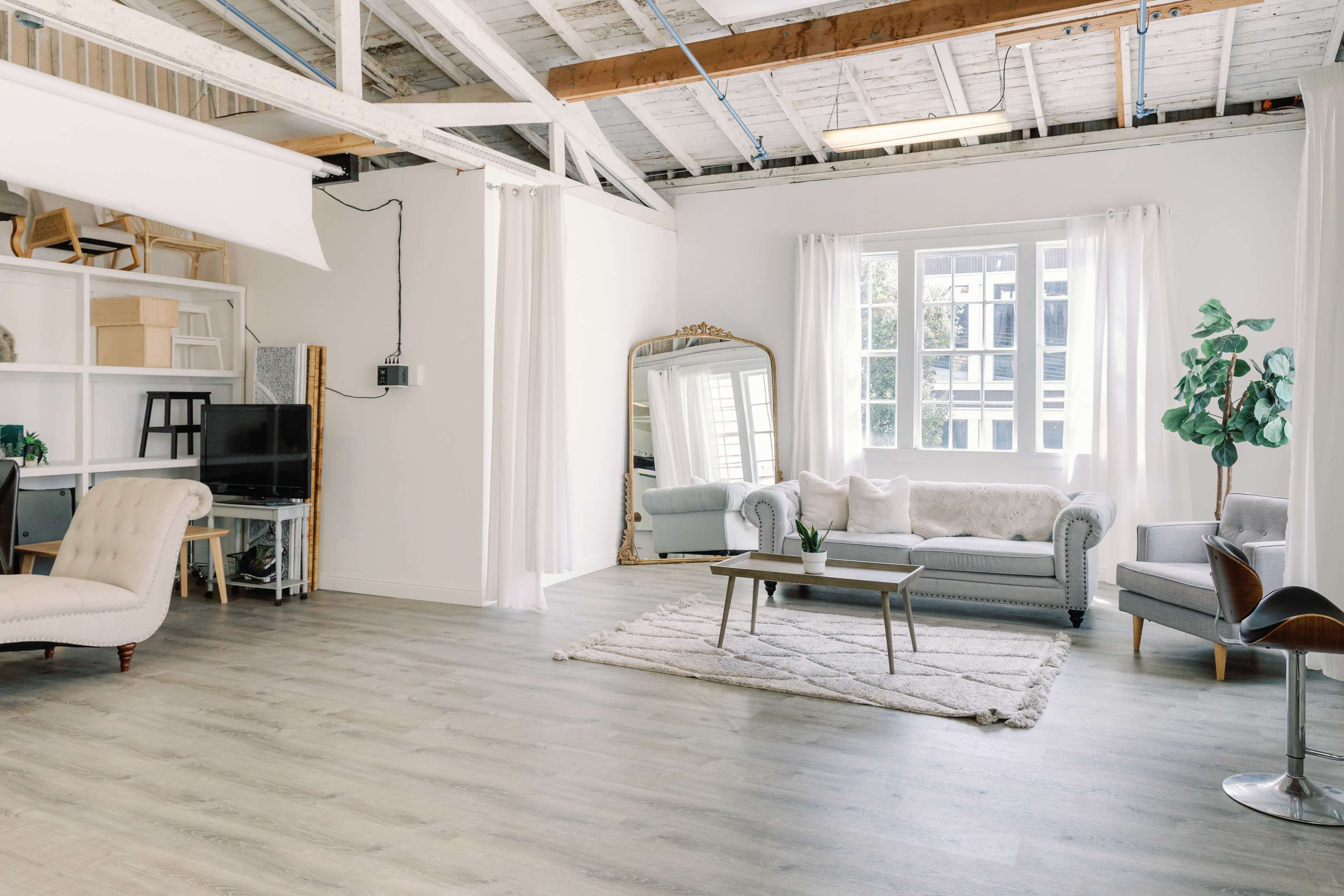 A spacious, bright room with a gray sofa, a coffee table, a large mirror, and plants, along with a work area featuring a computer and a chair.