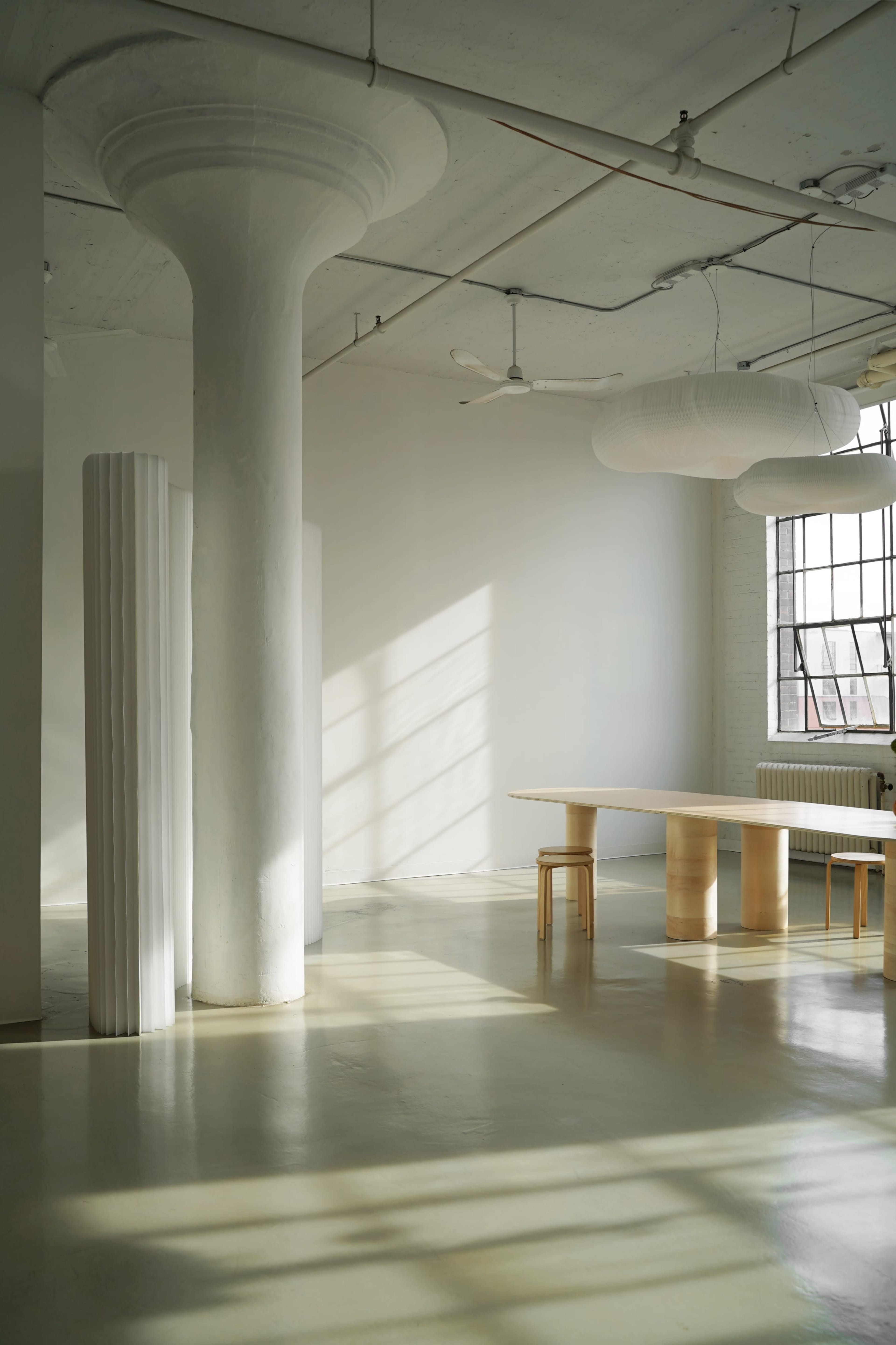A minimalist interior with a large wooden table and slender chairs beside tall white columns, illuminated by natural light from a large window.