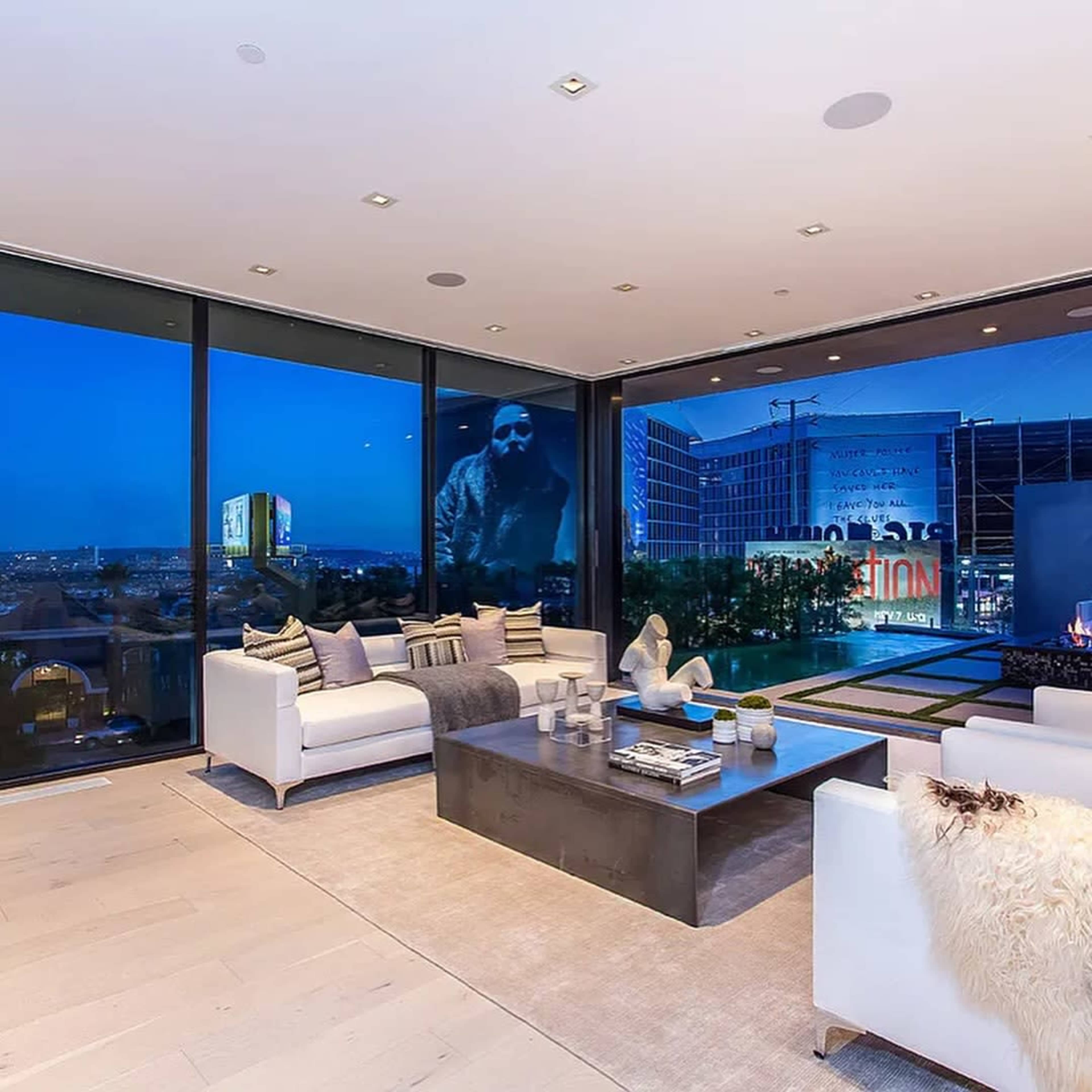 The modern living room features large windows overlooking a cityscape illuminated at dusk, with minimalist furniture and a sleek coffee table in the center.