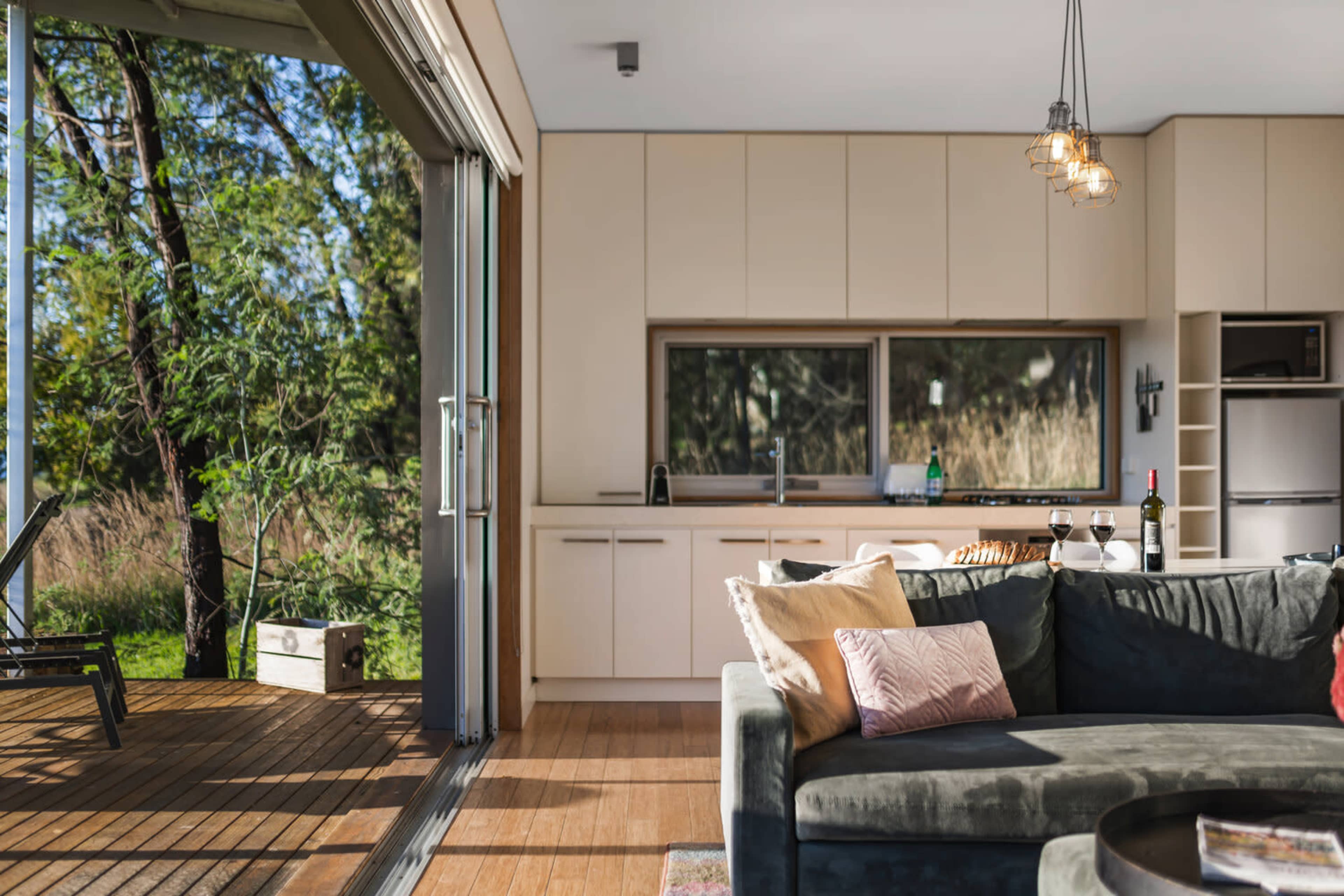 A modern kitchen and living area with large windows overlooking a wooden deck surrounded by greenery.