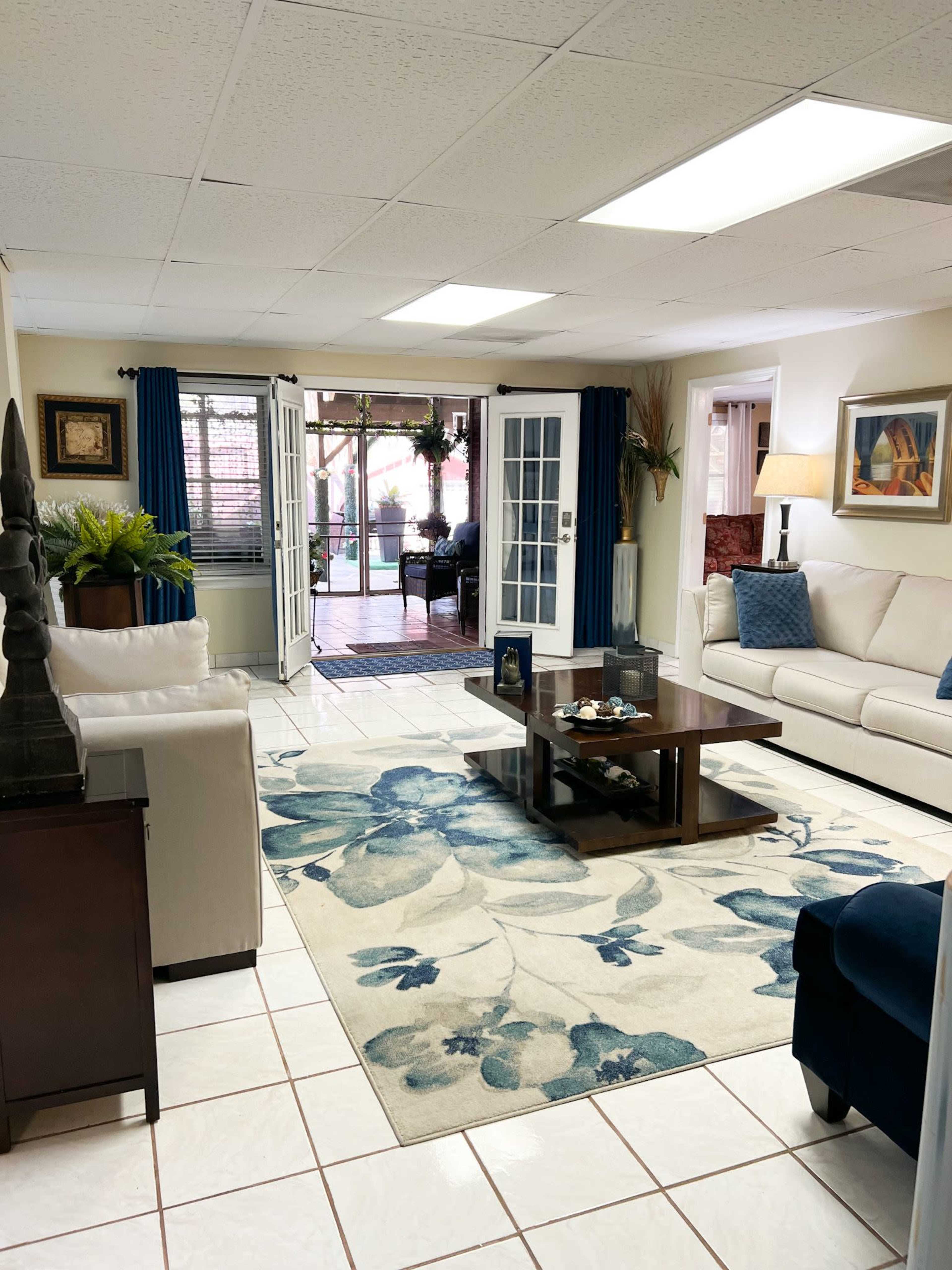A spacious living room with a white couch, a wooden coffee table, and a blue floral area rug, leading to a patio area visible through double French doors.