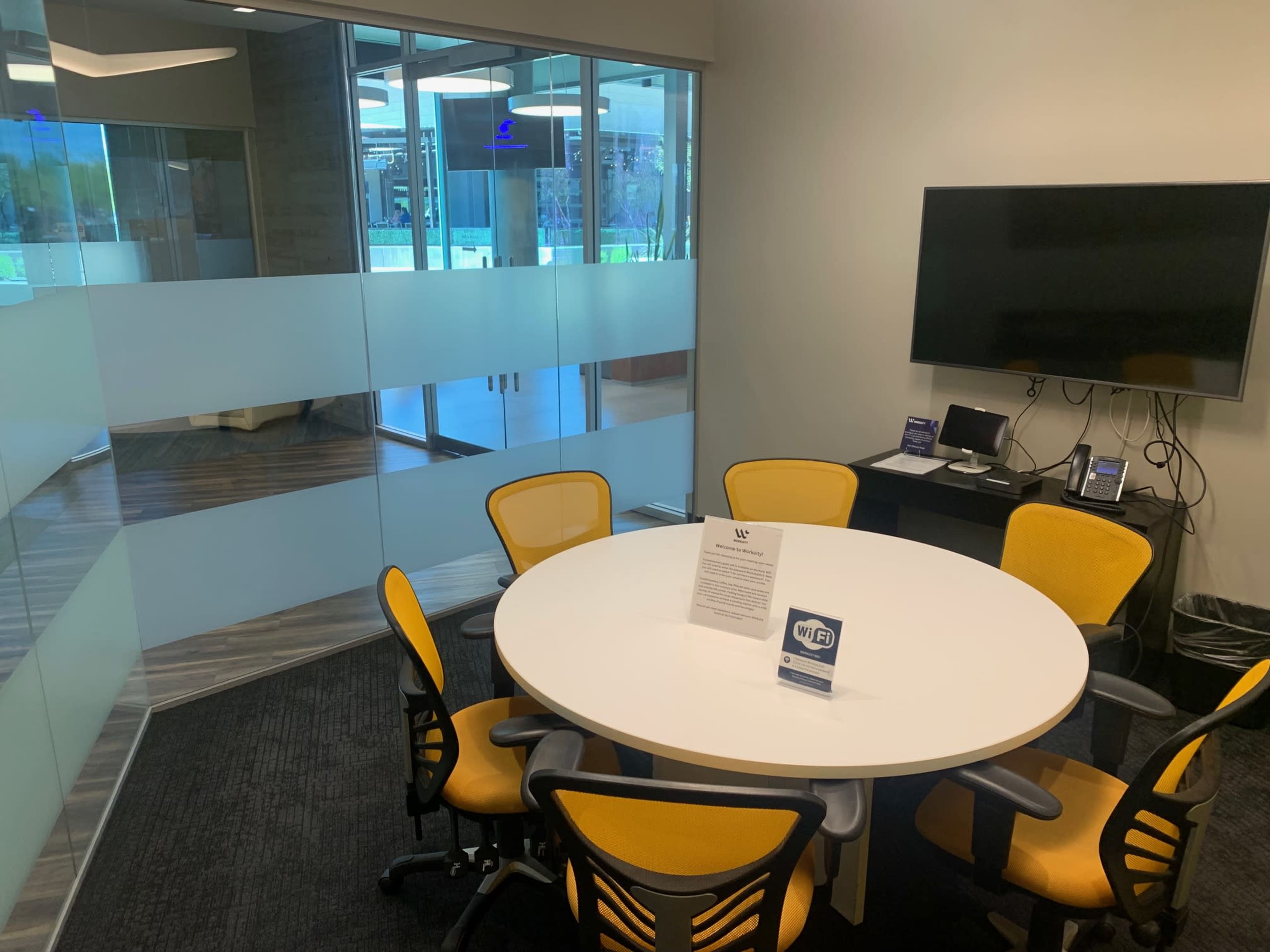 A round table with six yellow chairs is situated in a small meeting room featuring a wall-mounted television and glass partitions.