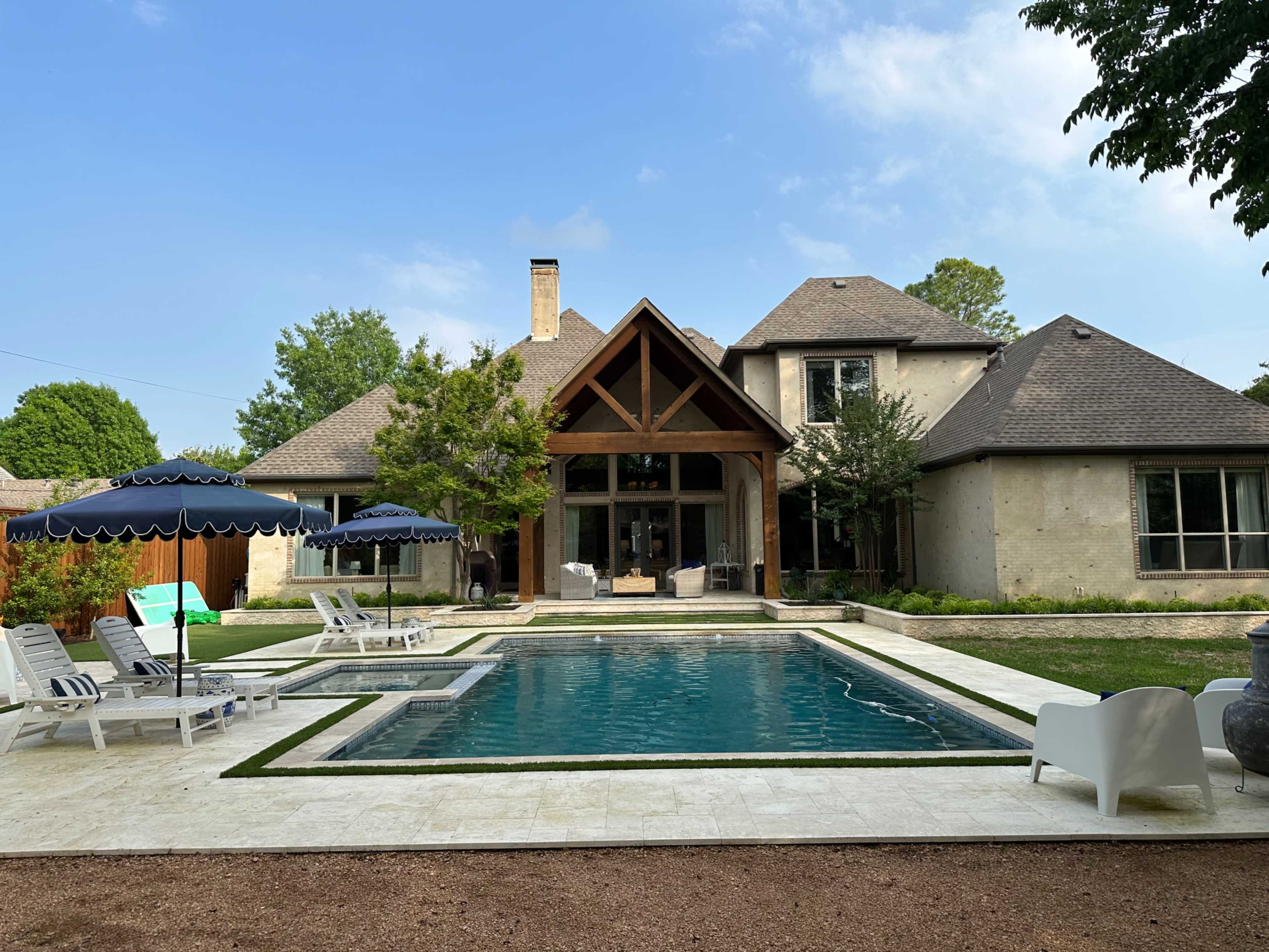 A modern house with a triangular roof and large windows is situated beside a rectangular swimming pool, surrounded by lounge chairs and umbrellas.