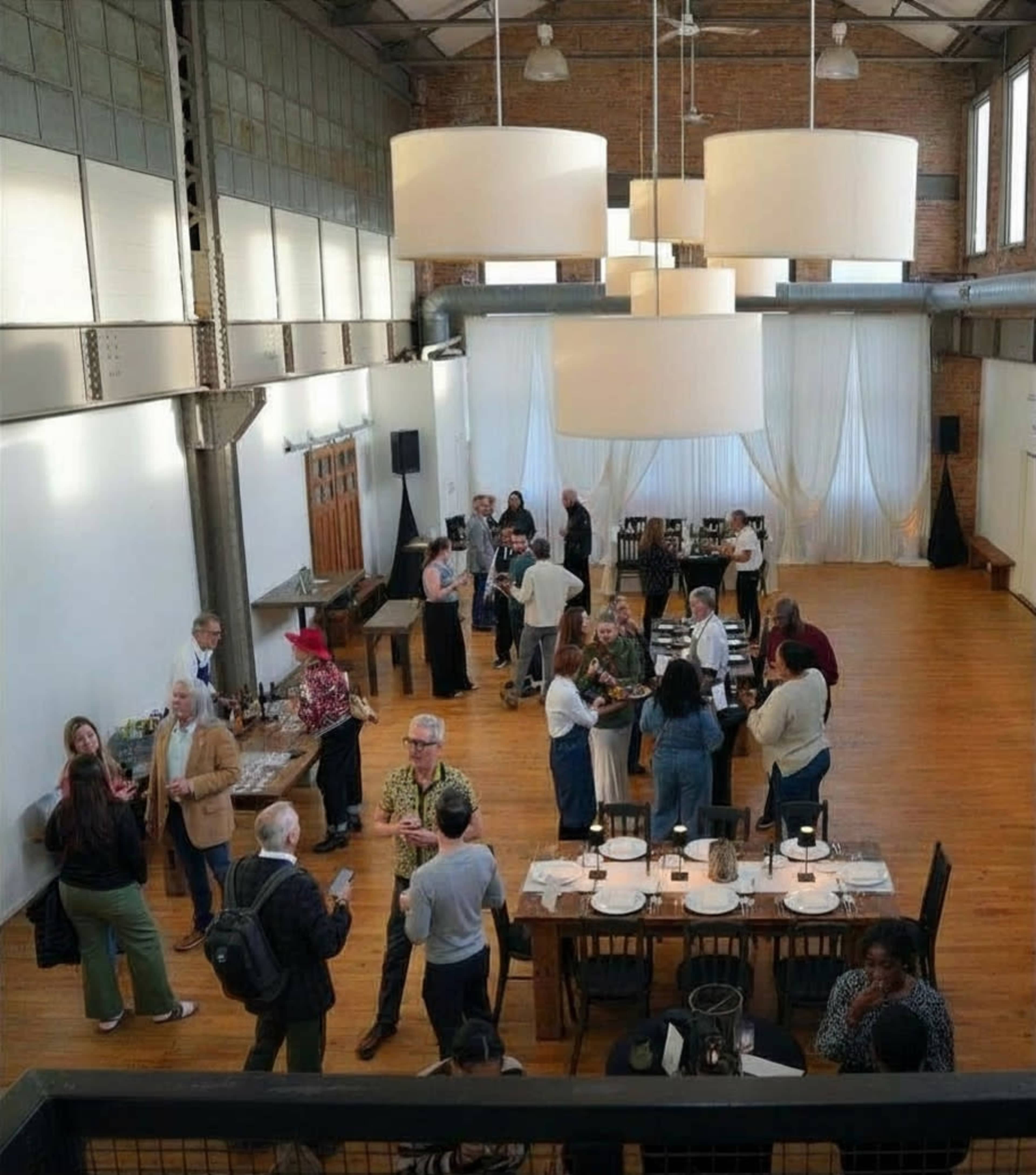 A spacious event venue features guests mingling at tables, with high ceilings and large pendant lights illuminating the scene.