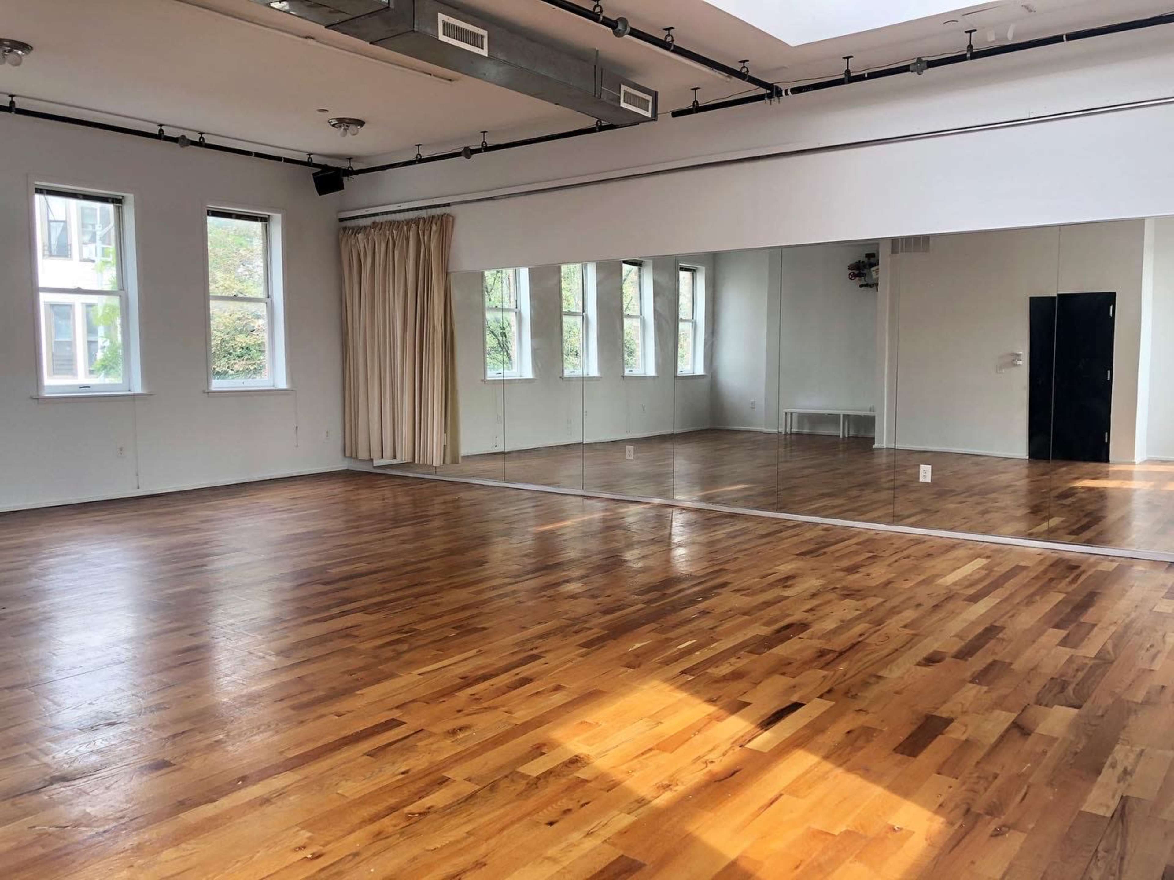 An empty room with wooden flooring, large windows, and a mirrored wall.